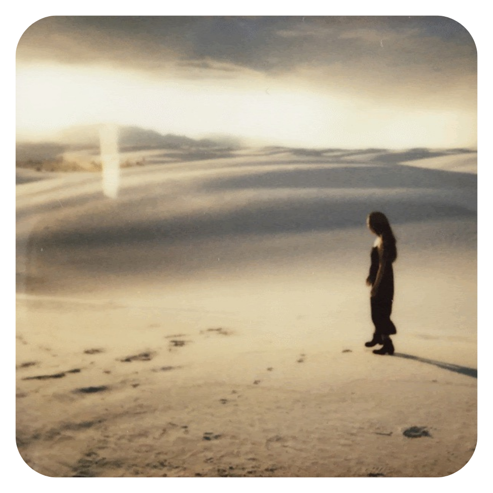 Heeya So standing at White Sands, New Mexico, looking ahead, with footprints in the sand. The sky is cloudy with a bright light source near the horizon.