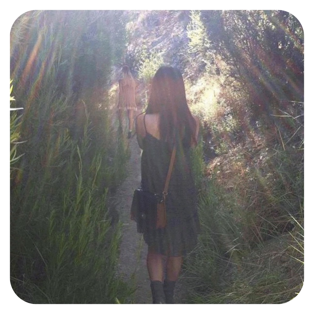 Heeya So walking along a narrow dirt trail surrounded by tall green grass and trees, with sunlight filtering through the foliage.