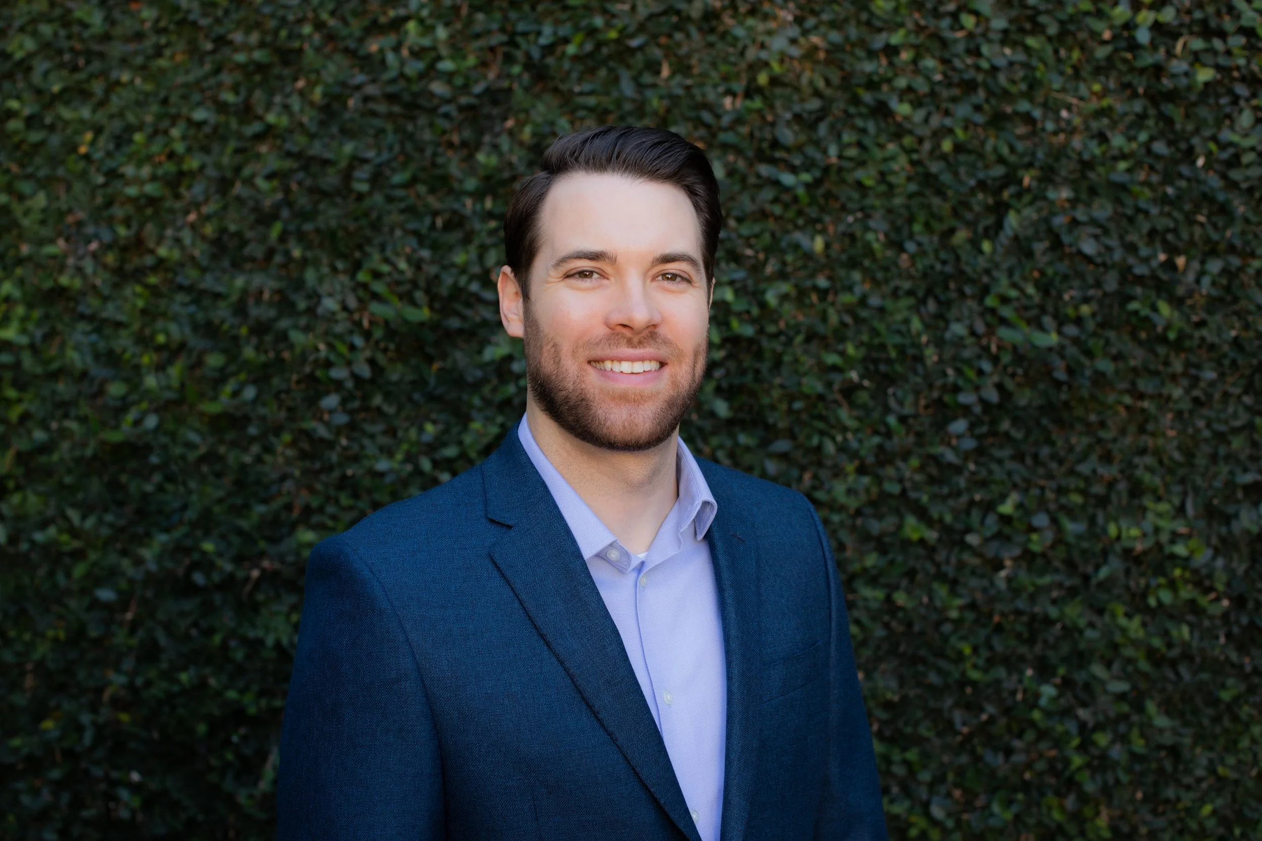A smiling man with dark hair and a beard wearing a dark blue blazer and light blue shirt stands in front of a green leafy hedge.