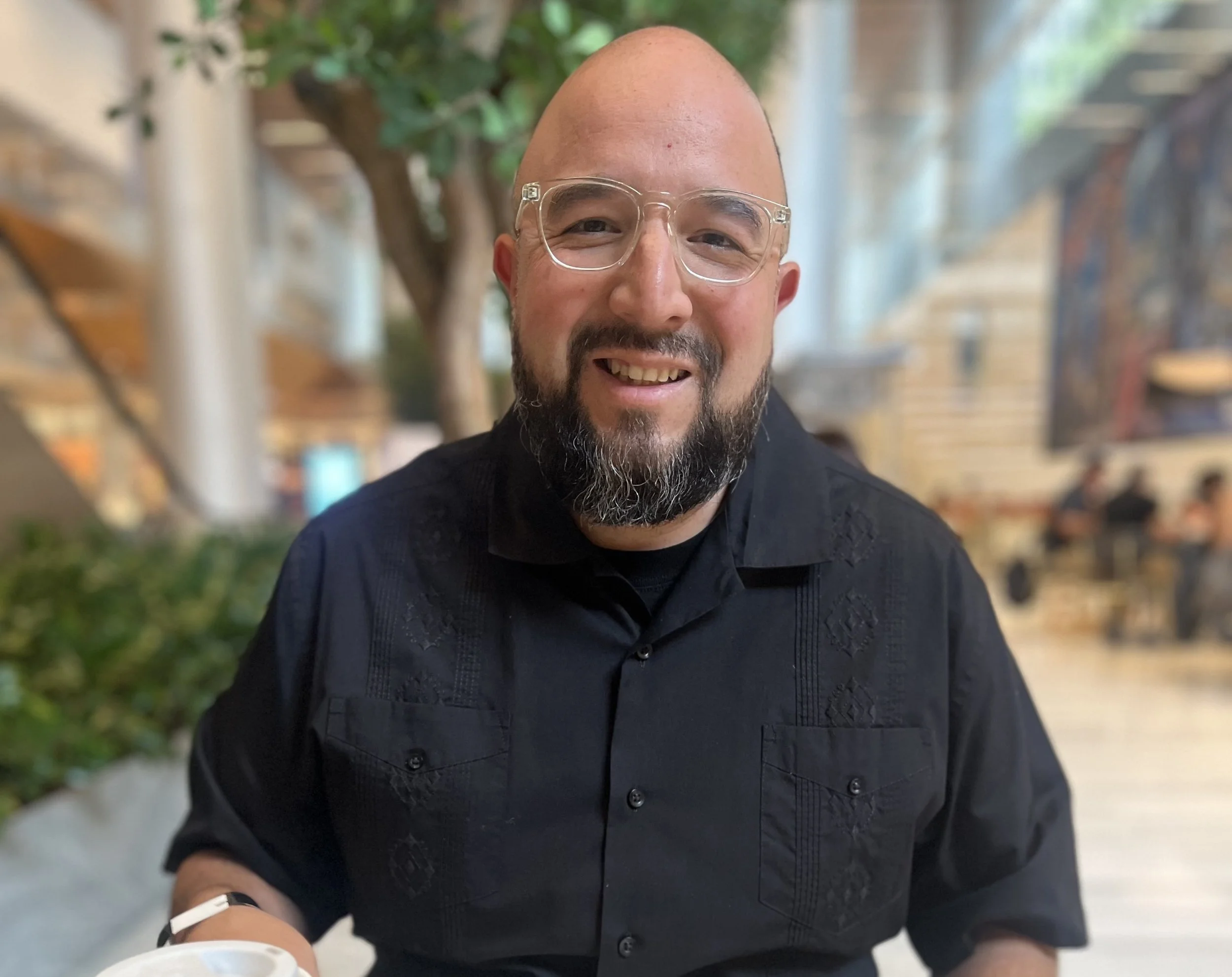 A smiling man with a beard, glasses, and a black shirt, sitting indoors with a blurred background of trees and people.