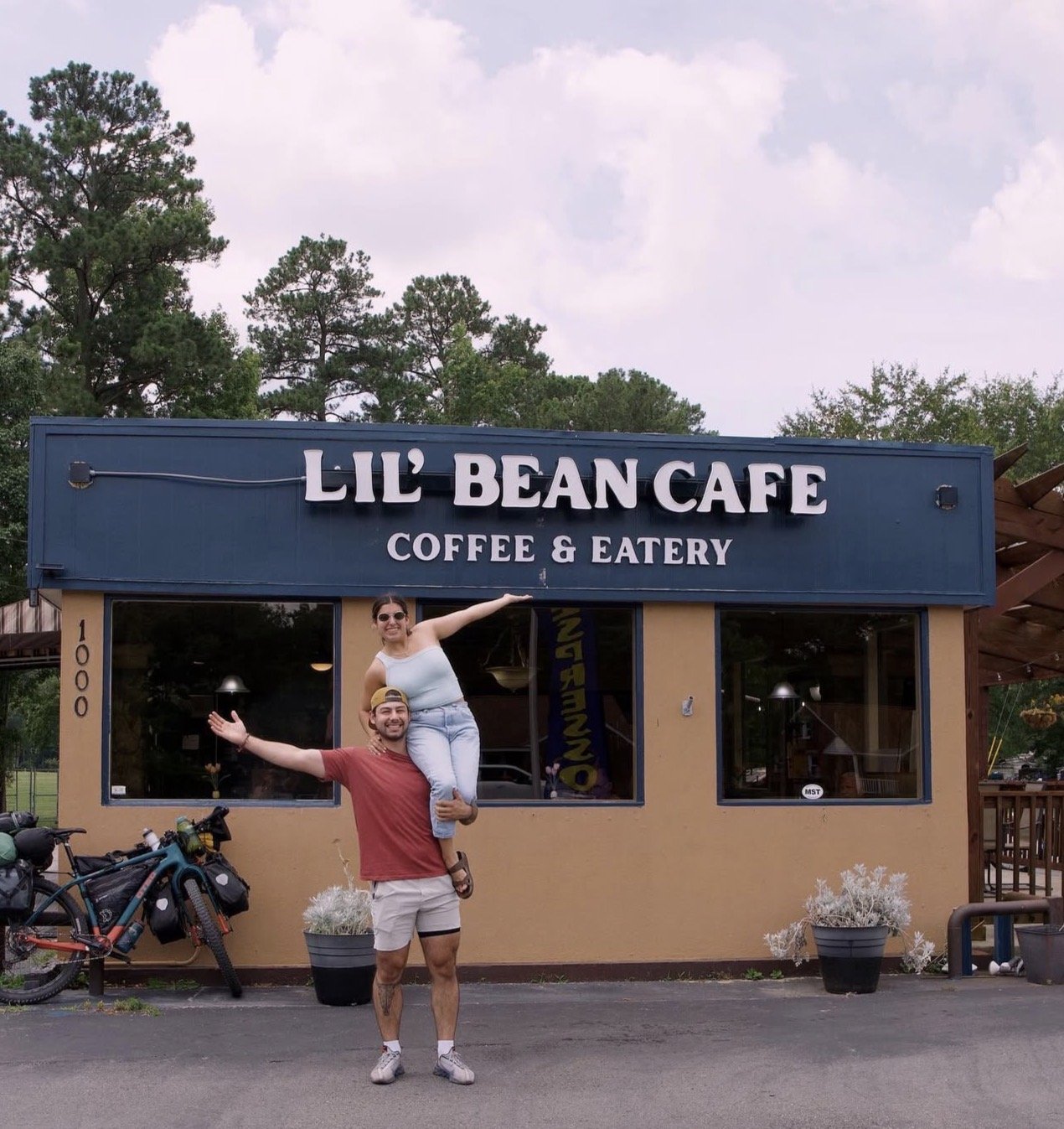 A man holding a woman on his shoulders in front of Lil' Bean Cafe, a coffee and eatery, with bicycles and potted plants nearby, and trees in the background.