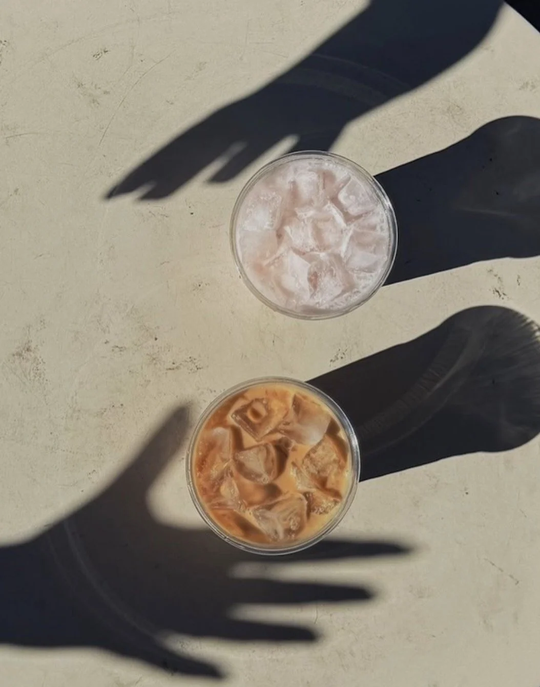 Two glasses of iced beverages, one with a pinkish drink and ice cubes, the other with a brownish drink and ice cubes, viewed from above on a light-colored surface. Shadows of three hands are cast on the surface.