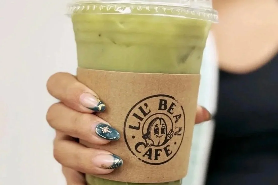 Hand holding a clear plastic cup with a green beverage and a clear plastic lid. The cup has a brown paper sleeve with the logo 'LIL' BEA CAFE' and a smiling character making a peace sign. The person's nails are decorated with nail art featuring stars and metallic accents.