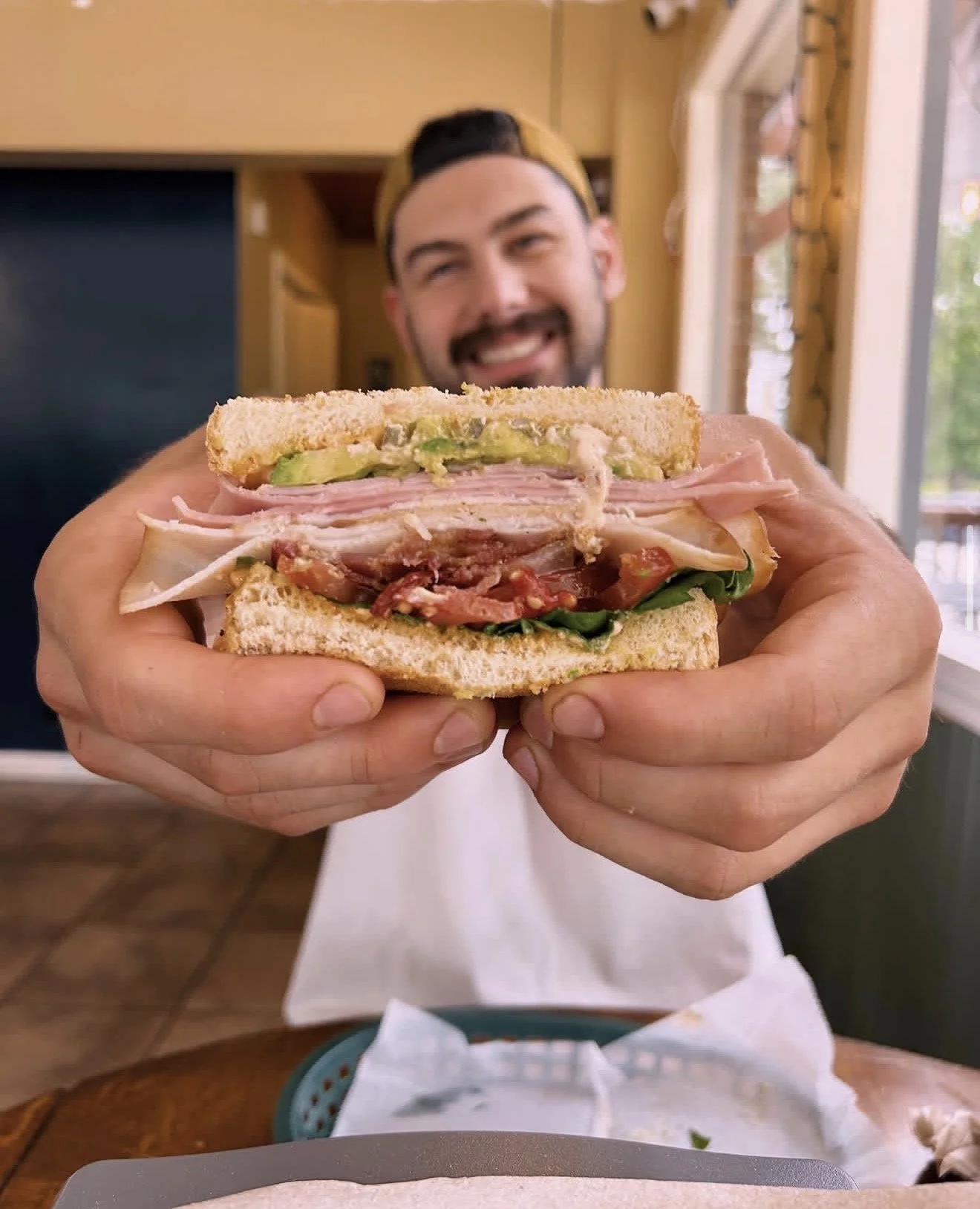 A person holding a sandwich with layers of ham, turkey, bacon, lettuce, tomato, and avocado in a sandwich bun, with a smiling man in the background.