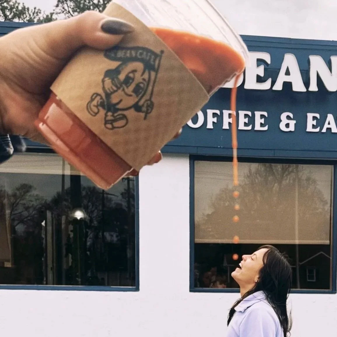 A woman standing outside a cafe, looking up with a smile, as a hand pours hot coffee from a cup with a cartoon mascot and the word 'delicious' on it, with the coffee streaming down.