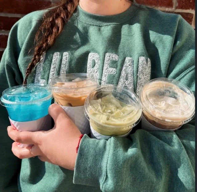 A person wearing a green sweatshirt holding four cups of colorful ice cream or frozen yogurt with clear lids, standing outdoors against a brick wall.