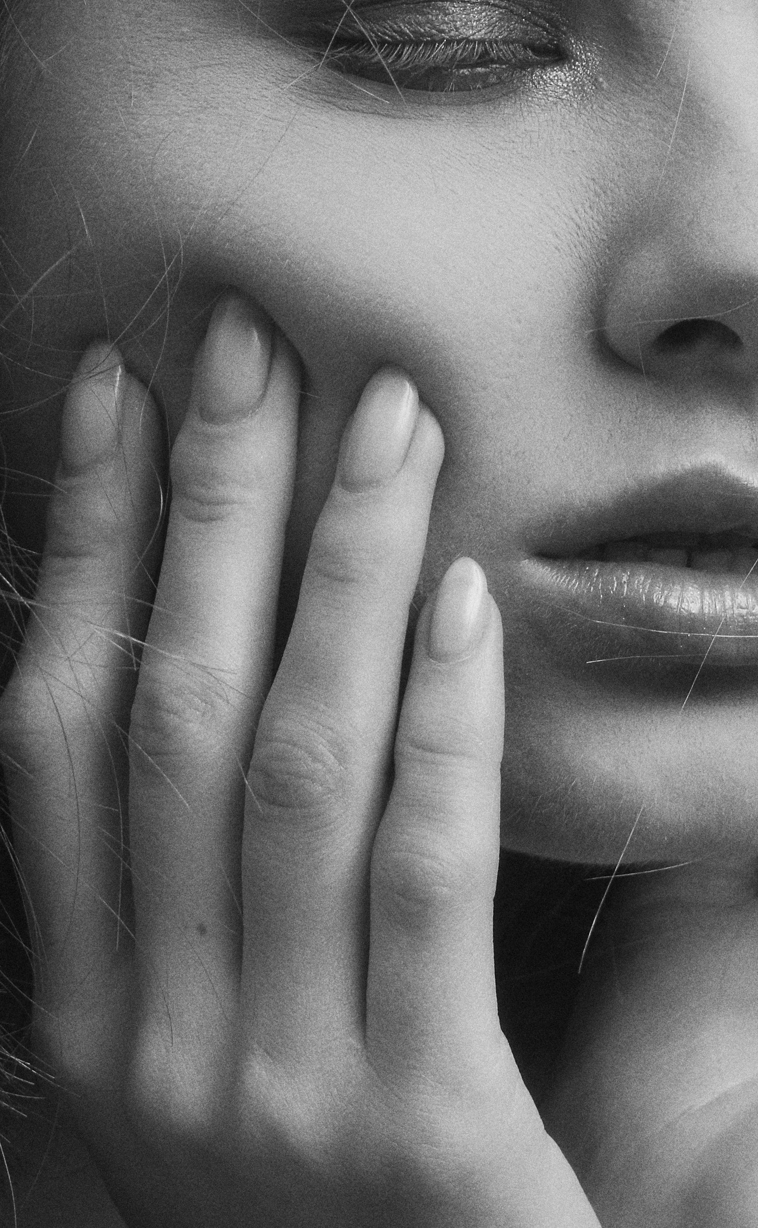 A close-up black and white photo of a woman's face, with her hand partially covering her cheek and lips. Only part of her face, eye, nose, and lips are visible.