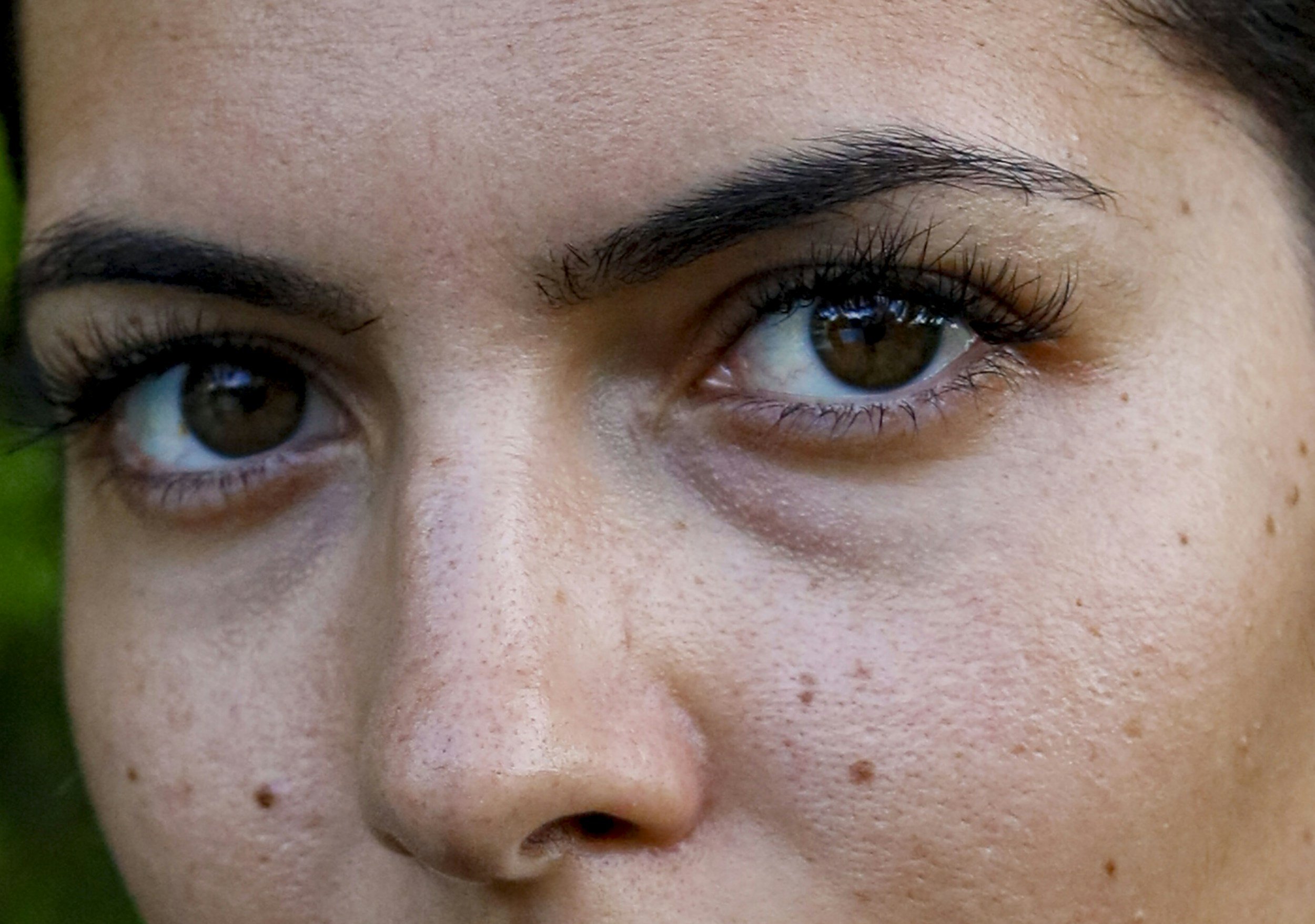 Close-up of a person's face showing brown eyes, dark eyebrows, and freckles, with some green foliage visible on the left side.