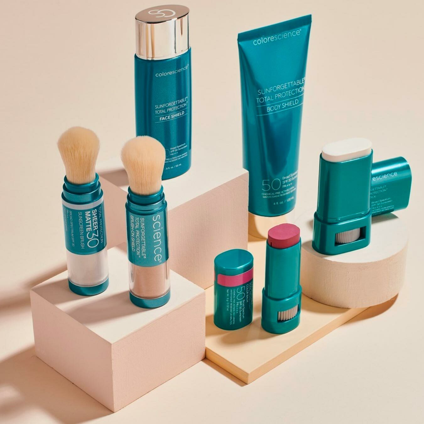A collection of Colorescience skincare and makeup products arranged on pastel-colored platforms, including sunscreen sticks, a face shield spray, and a body shield tube, with two makeup brushes.
