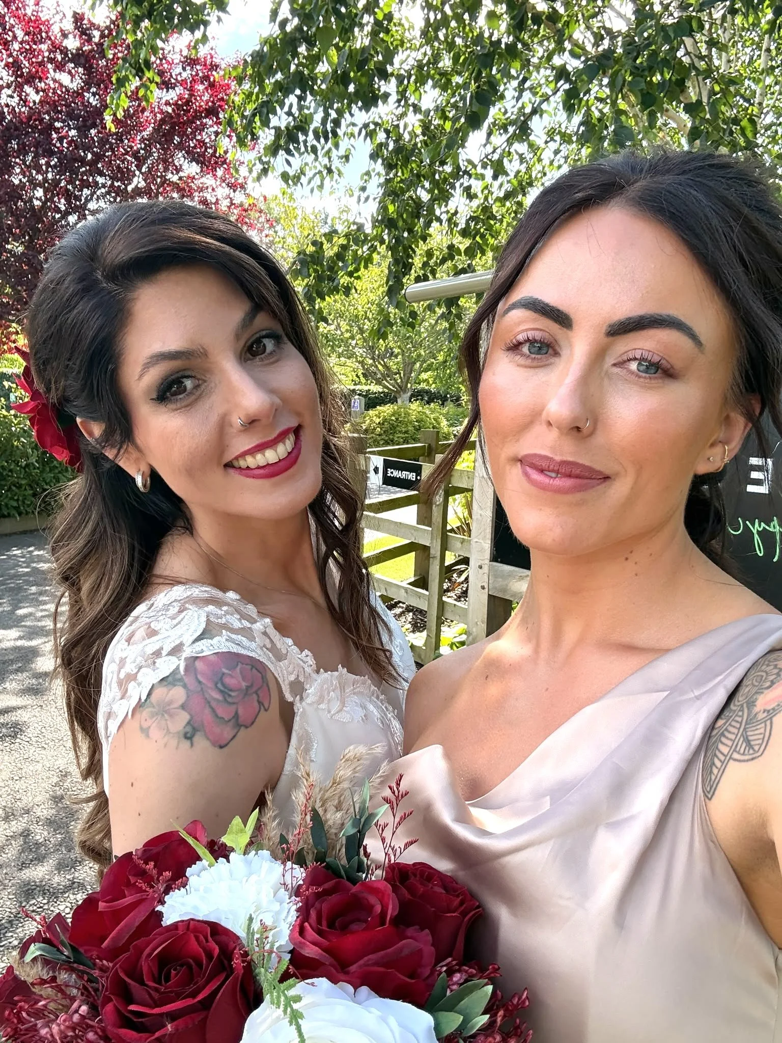 Two women on their wedding day, standing outdoors with trees and bright sunlight behind them. The woman on the left has dark brown hair, a floral hair accessory, and a lace wedding dress. The woman on the right has black hair, blue eyes, and is weari
