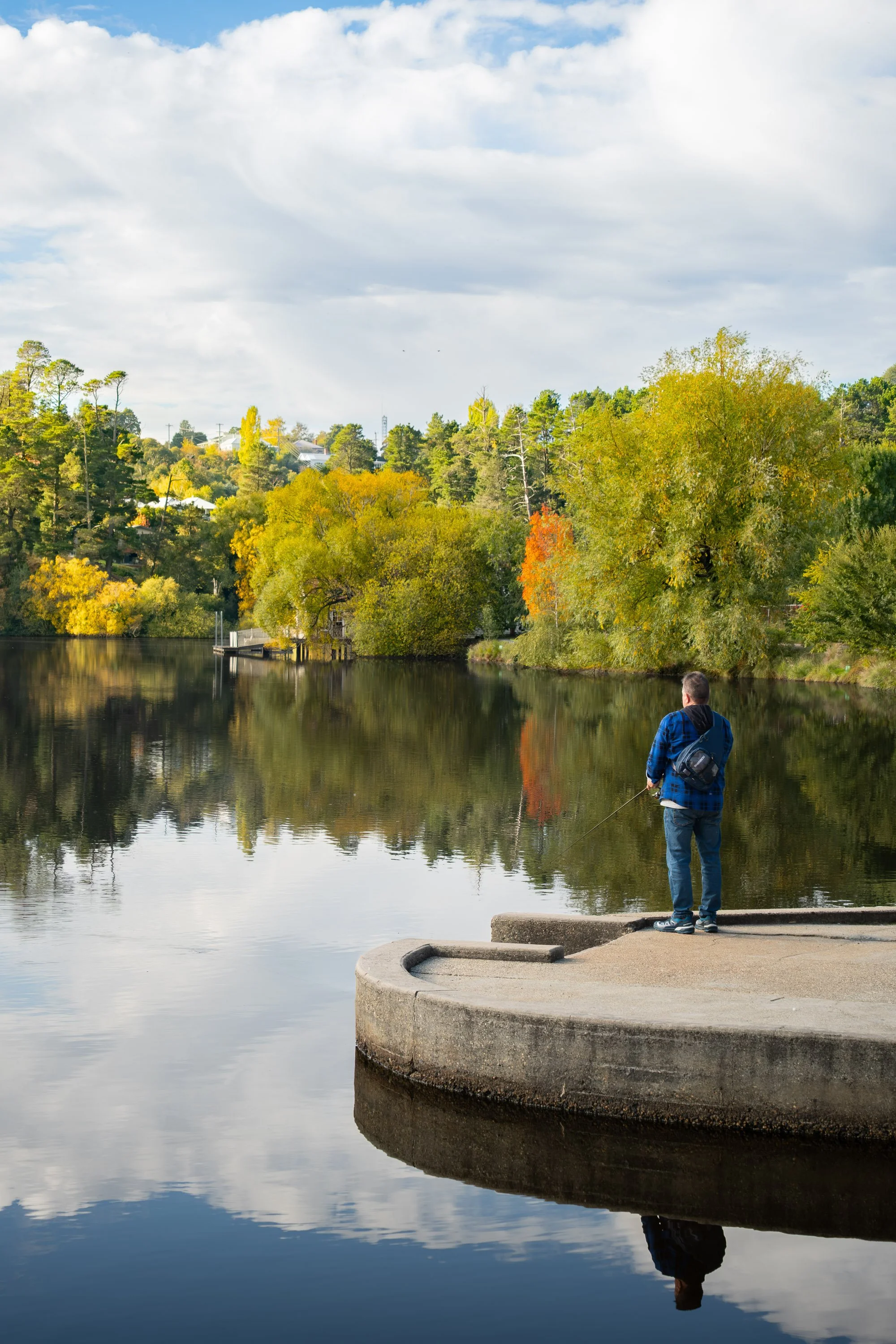 A man fishing on a concrete dock by a calm lake, with a background of trees with fall foliage and a cloudy sky reflected in the water.