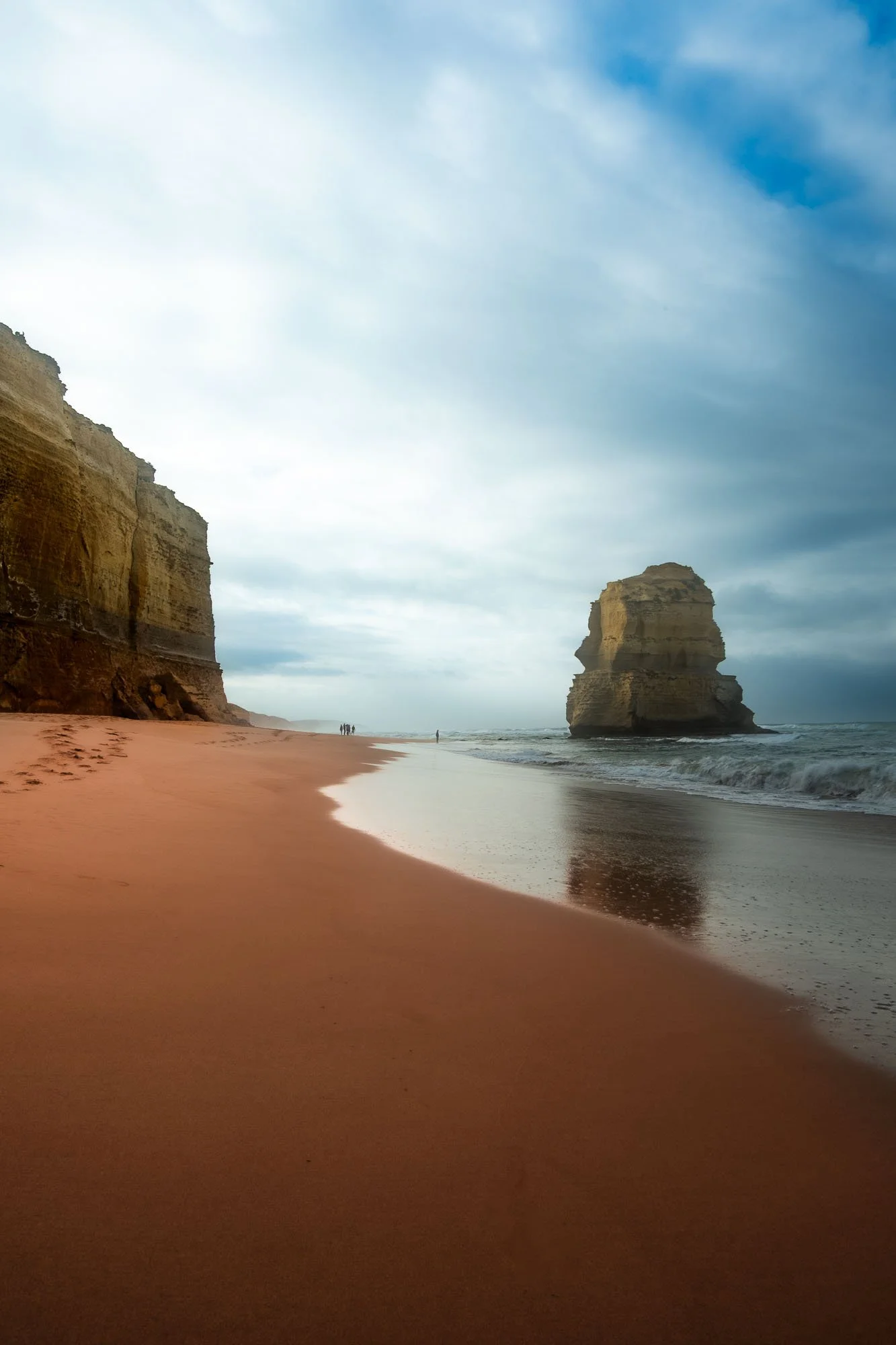 A beach with pink sand, white waves, towering sandy cliffs on the left, and a large sea stack formation in the ocean on the right, under a partly cloudy sky.