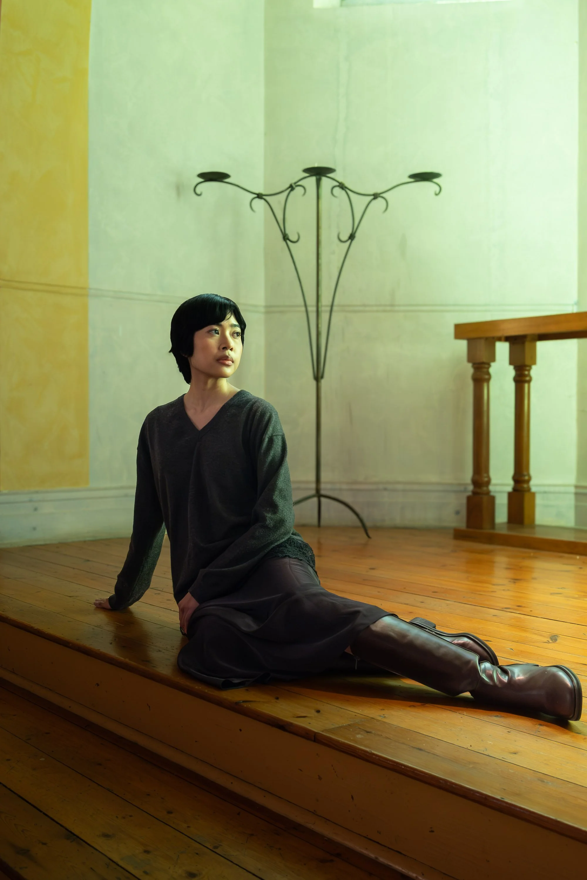 A woman with short black hair and a dark top sitting on a wooden stage floor, with one leg extended and the other bent, in a room with pale yellow and green walls and a wooden railing, with a metal floor lamp in the background.