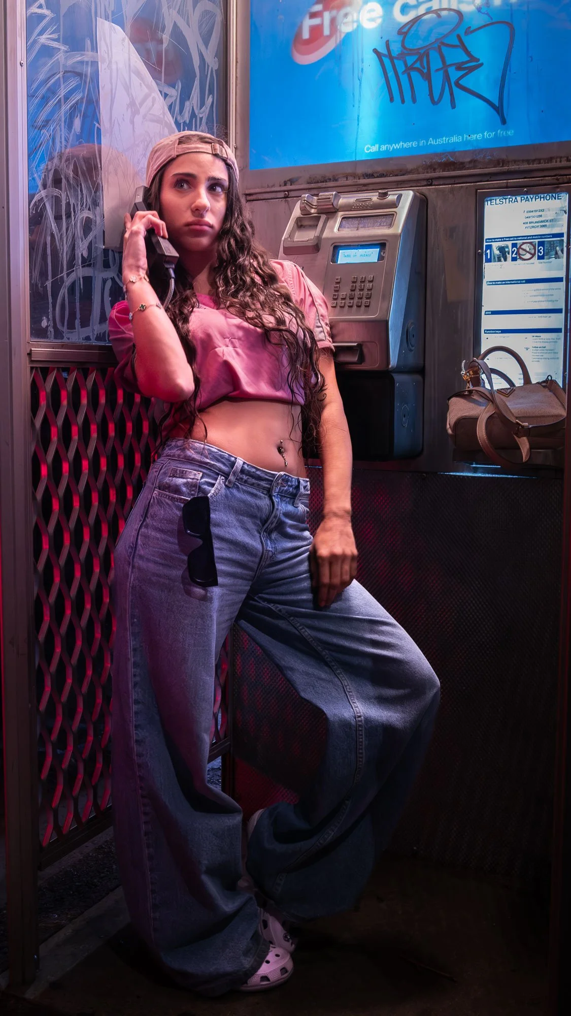Young woman with long curly hair, wearing a pink crop top, baggy jeans, and a baseball cap backwards, standing at a payphone booth inside a city at night.