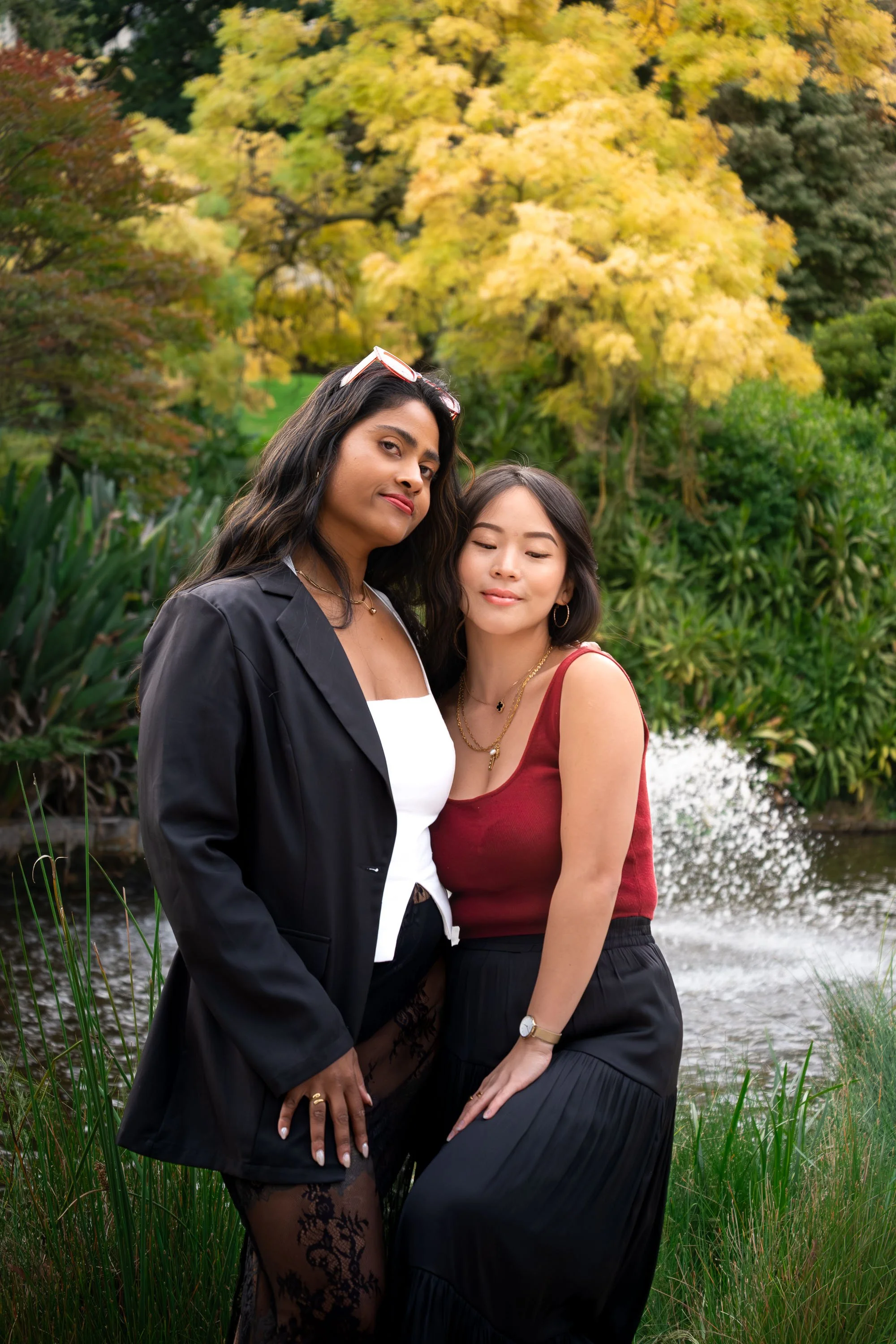 Two women standing outdoors near a body of water with greenery and autumn trees in the background. One woman has dark wavy hair, wearing a black blazer and lace stockings, and the other has straight dark hair, wearing a red sleeveless top and black skirt.