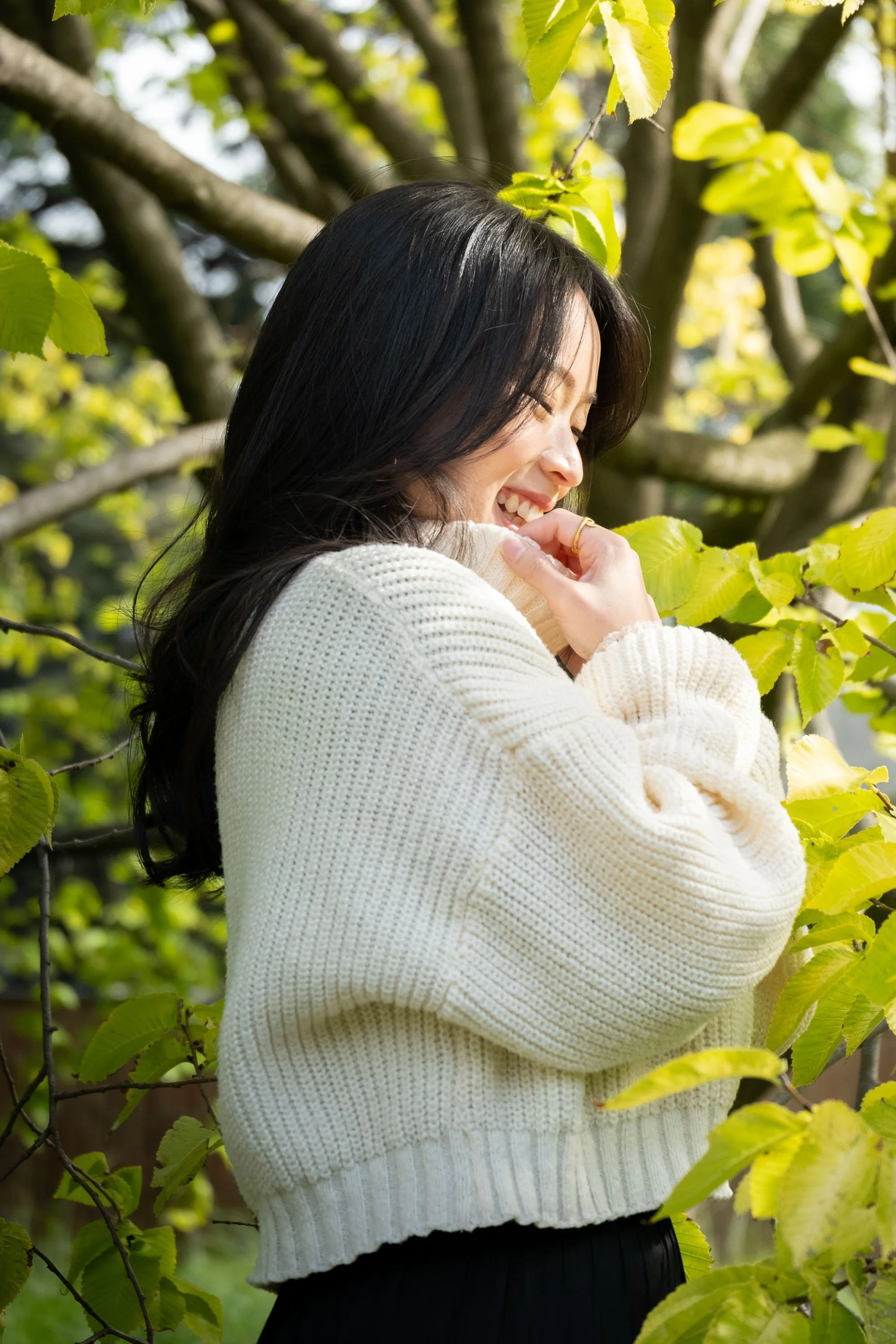 A woman with long dark hair smiling and covering her mouth with a hand, standing among green leaves in a natural outdoor setting.