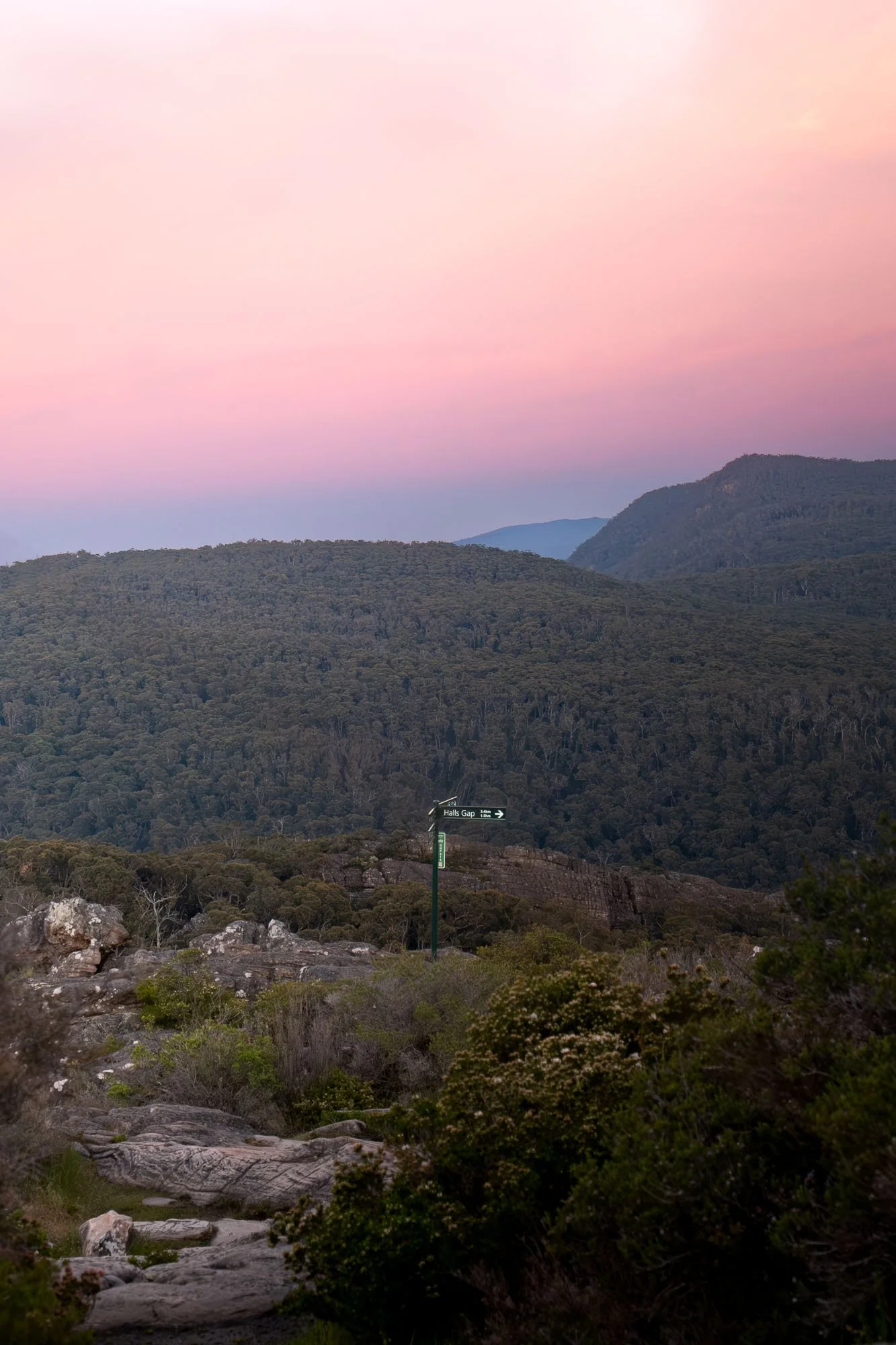 A scenic view of mountains covered with dense greenery at sunset or sunrise, with a pink and purple sky above and a small path with a signpost that reads "Halls Gap" in the foreground.
