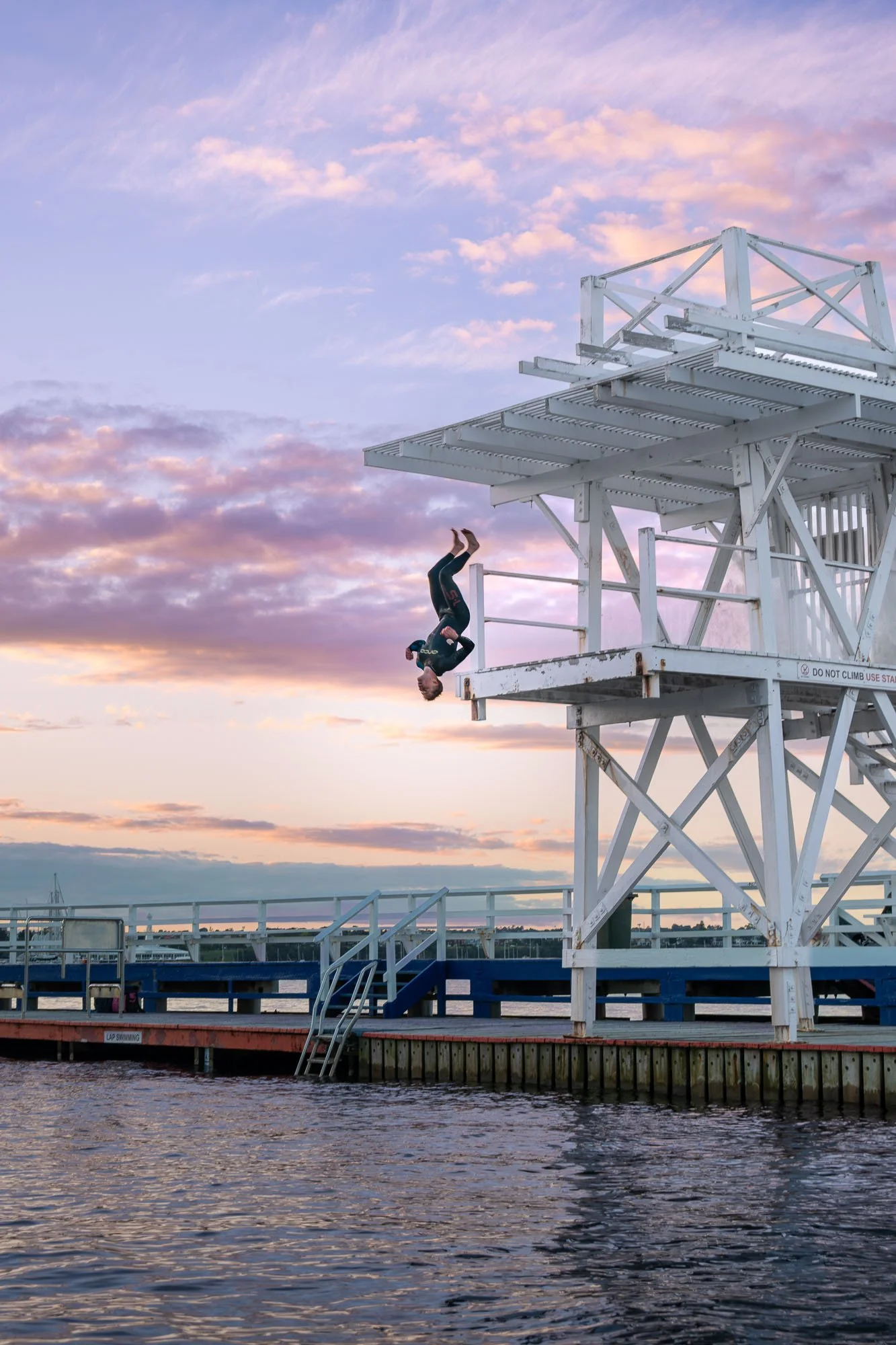A woman dressed in black performing a backflip off a white lifeguard tower at a pier during sunset, with a cloudy sky and water below.