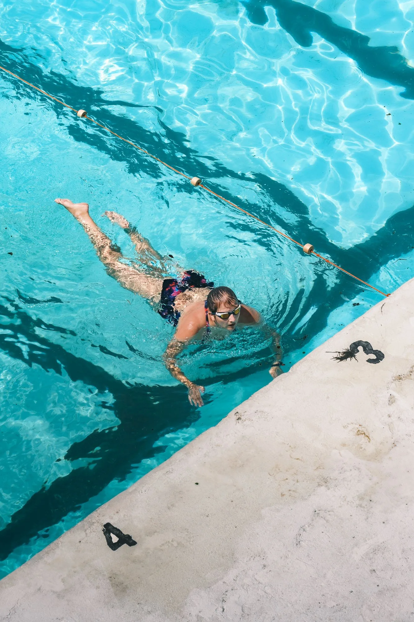 A person swimming in a pool near the edge, wearing goggles and swimwear, with a swimming lane marker floating in the water.