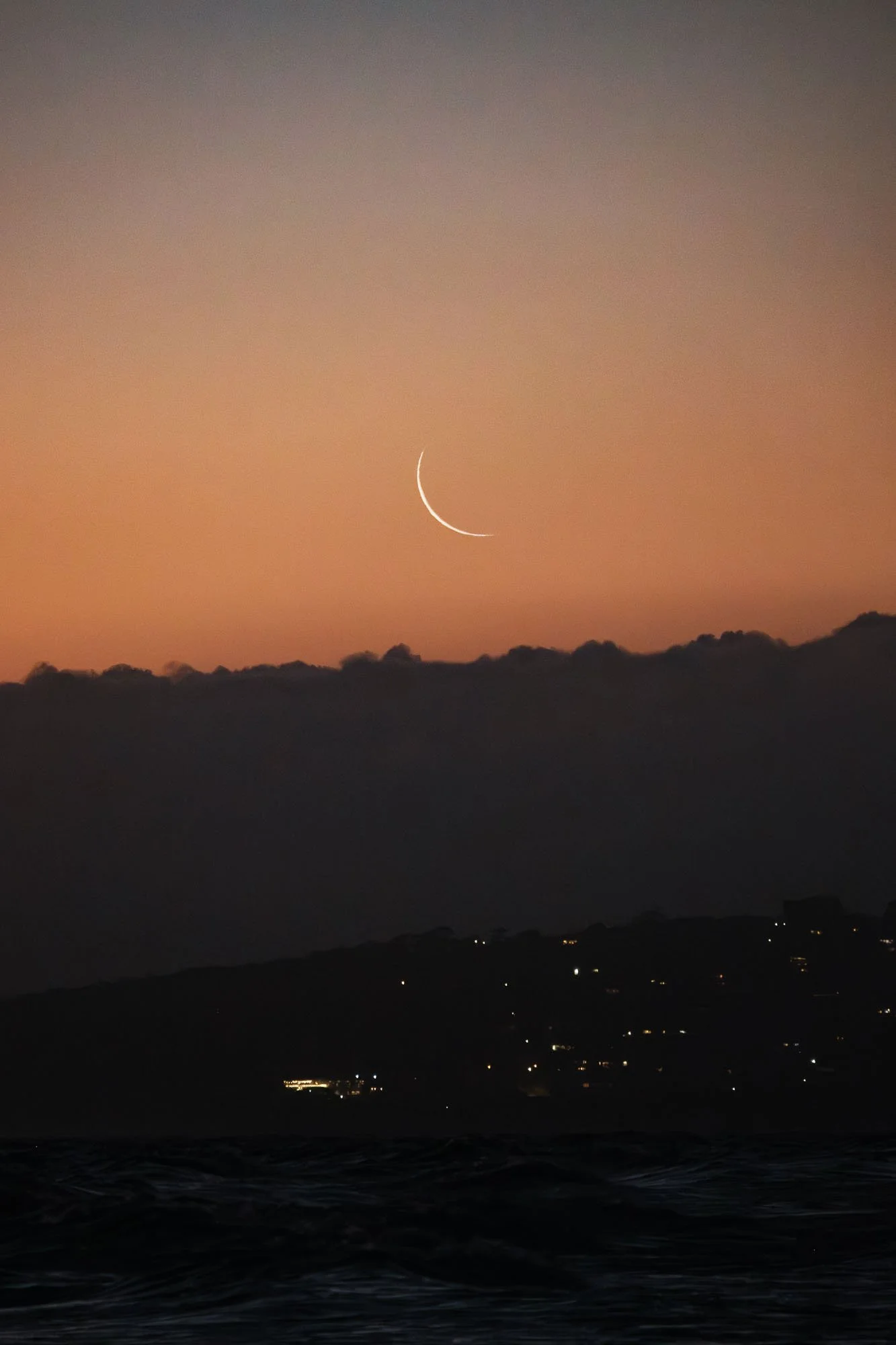 A photograph of a crescent moon in the sky during sunset or sunrise, with mountains and some scattered lights visible in the distance.