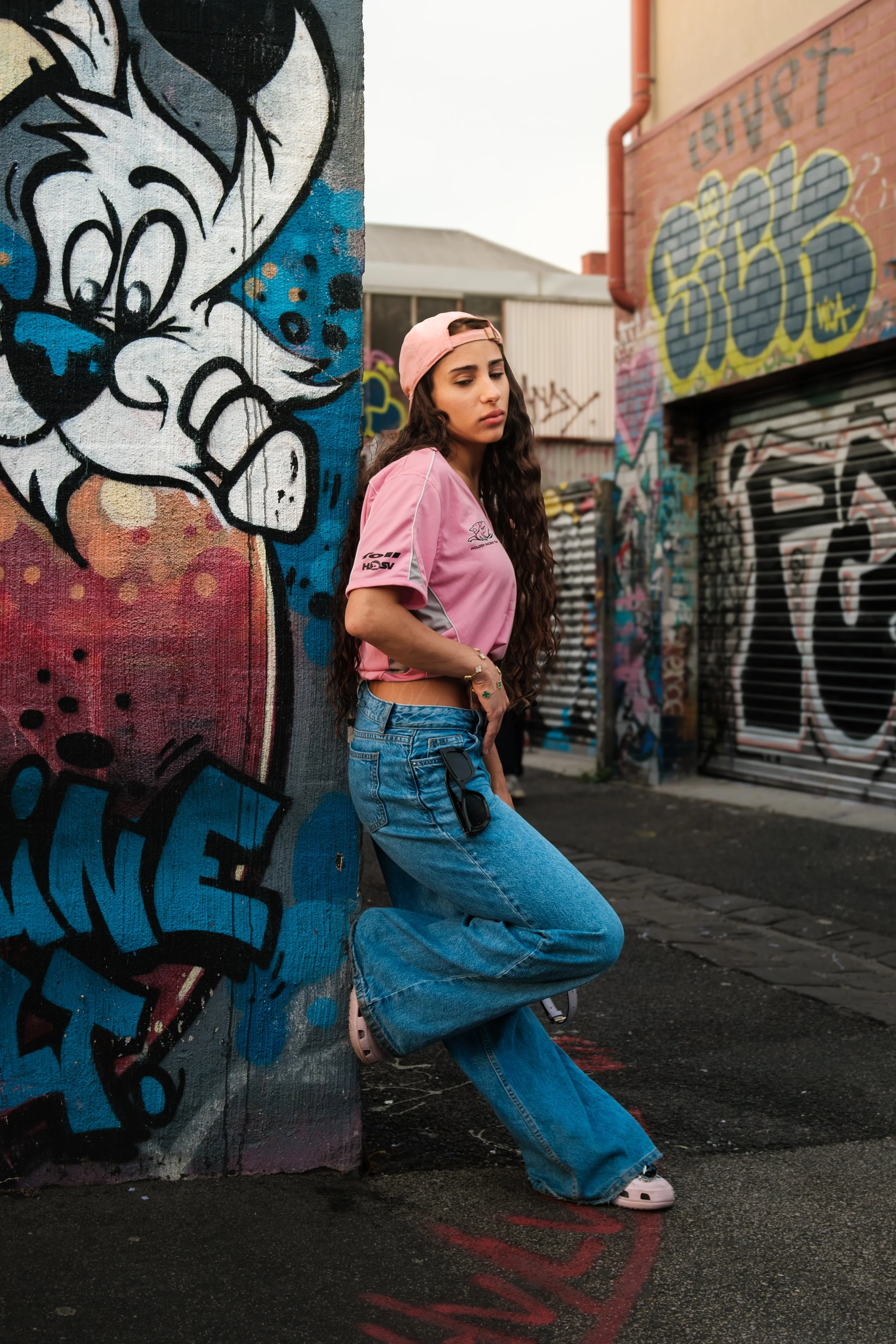 A young woman with long curly hair, wearing a pink cap, pink T-shirt, and blue jeans, leaning against a graffiti-covered wall in an urban alley. She has a contemplative expression, with her eyes closed and one hand in her pocket. The graffiti on the 