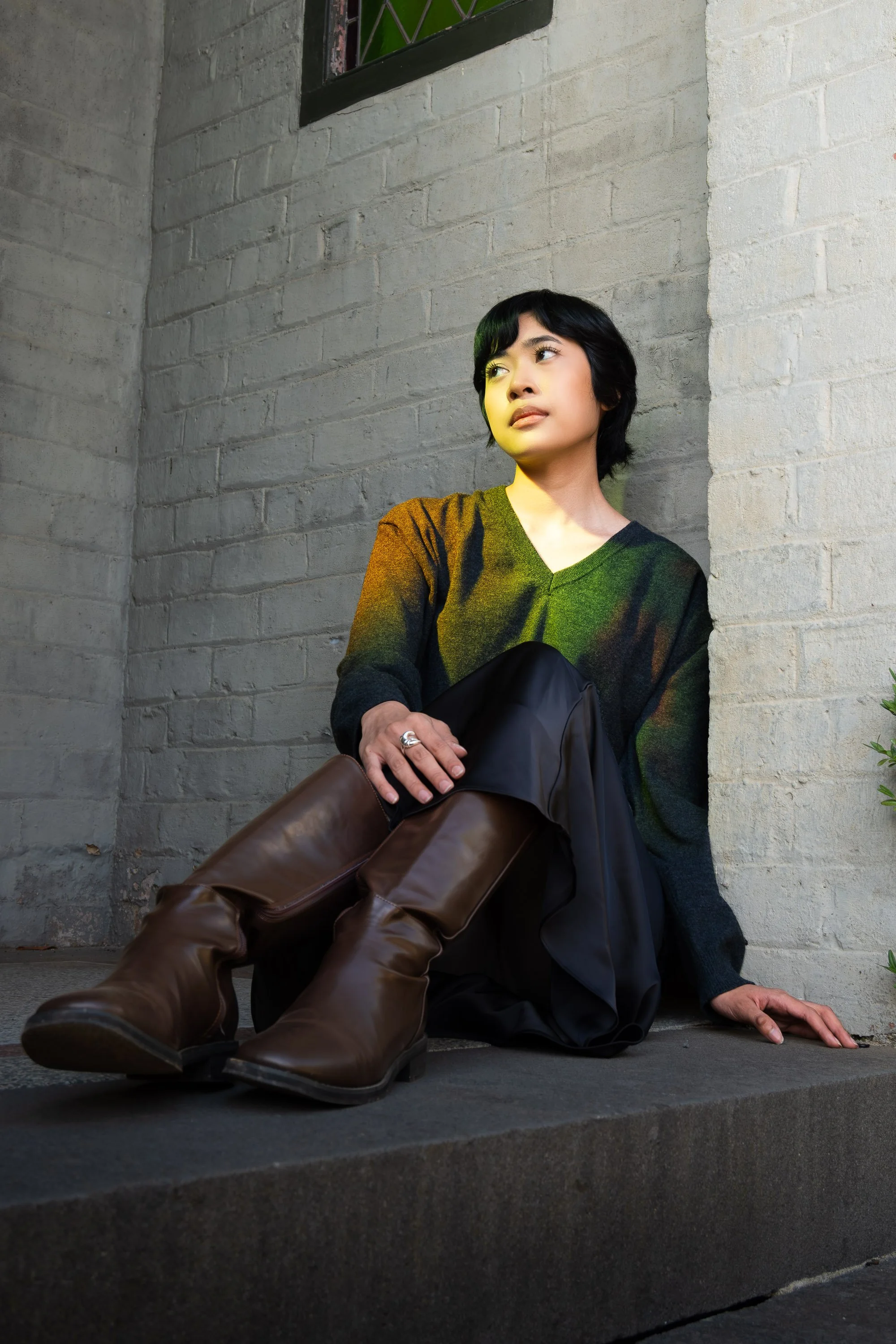 A young woman with short black hair, wearing a dark sweater, is sitting against a brick wall, looking thoughtfully into the distance. She has a silver ring on her left hand, which rests on her knee, and a pair of brown boots and black pants are visib