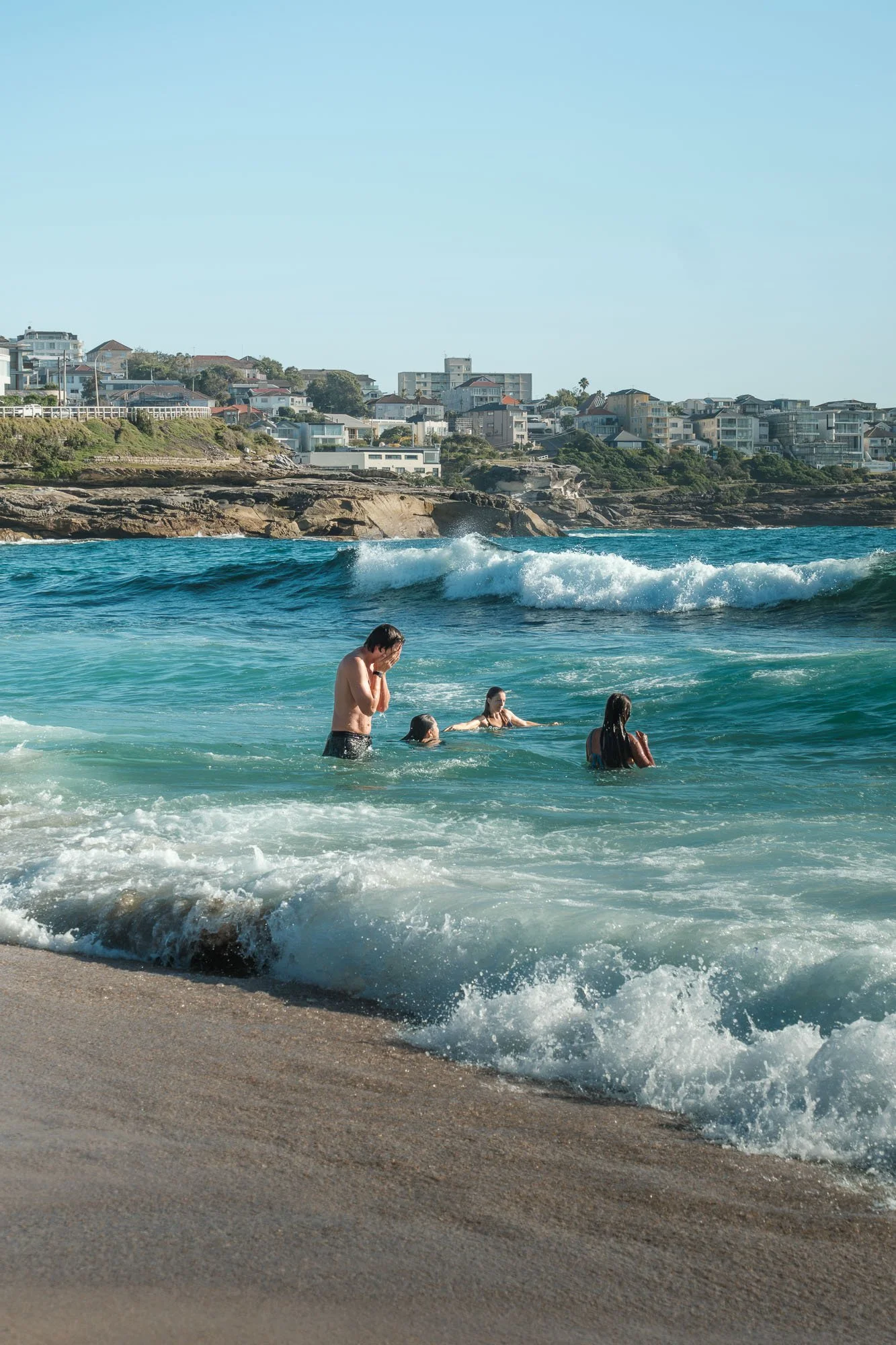 People enjoying a day at the beach, with some wading in the water and a cityscape of houses and buildings on a hillside in the background.