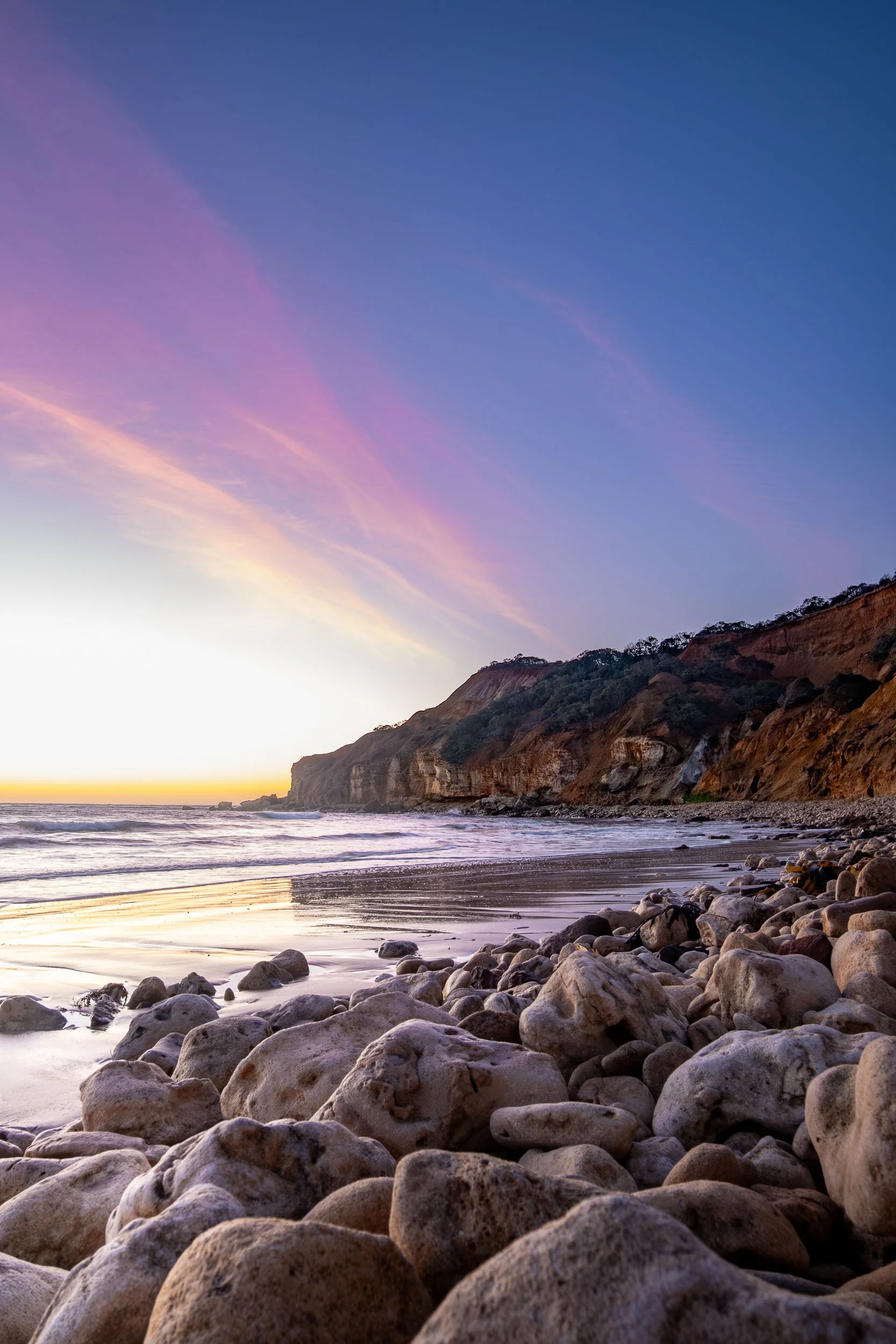 A rocky shoreline at sunset with cliffs in the distance, colorful sky with pink and blue clouds, and gentle waves lapping against the rocks.