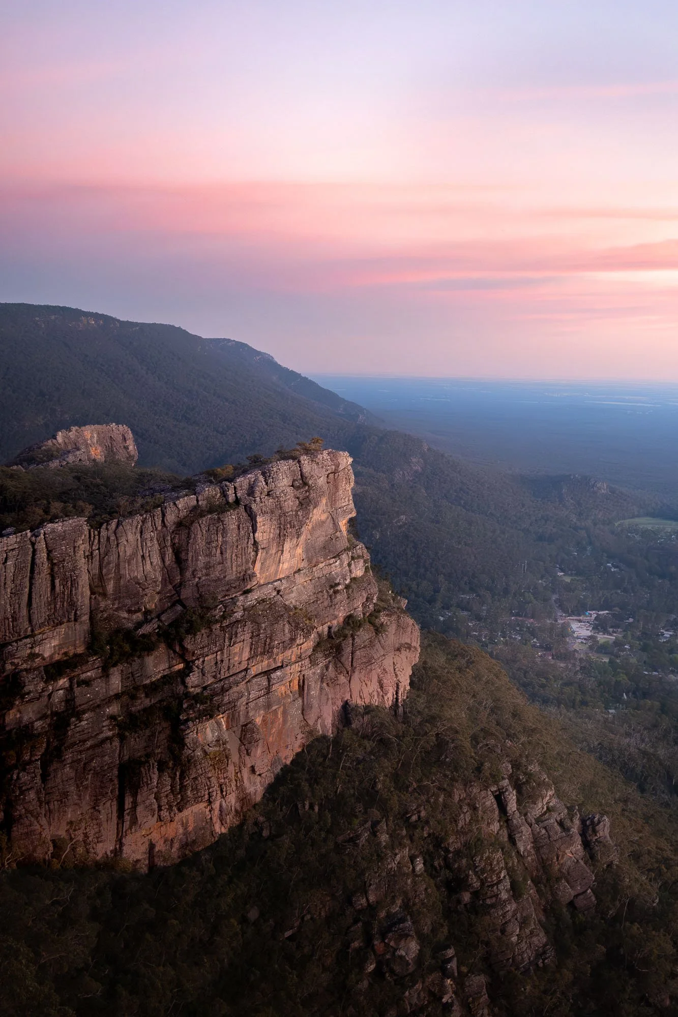Scenic view of a mountain cliff with forest below and a pastel-colored sky at sunset or sunrise.
