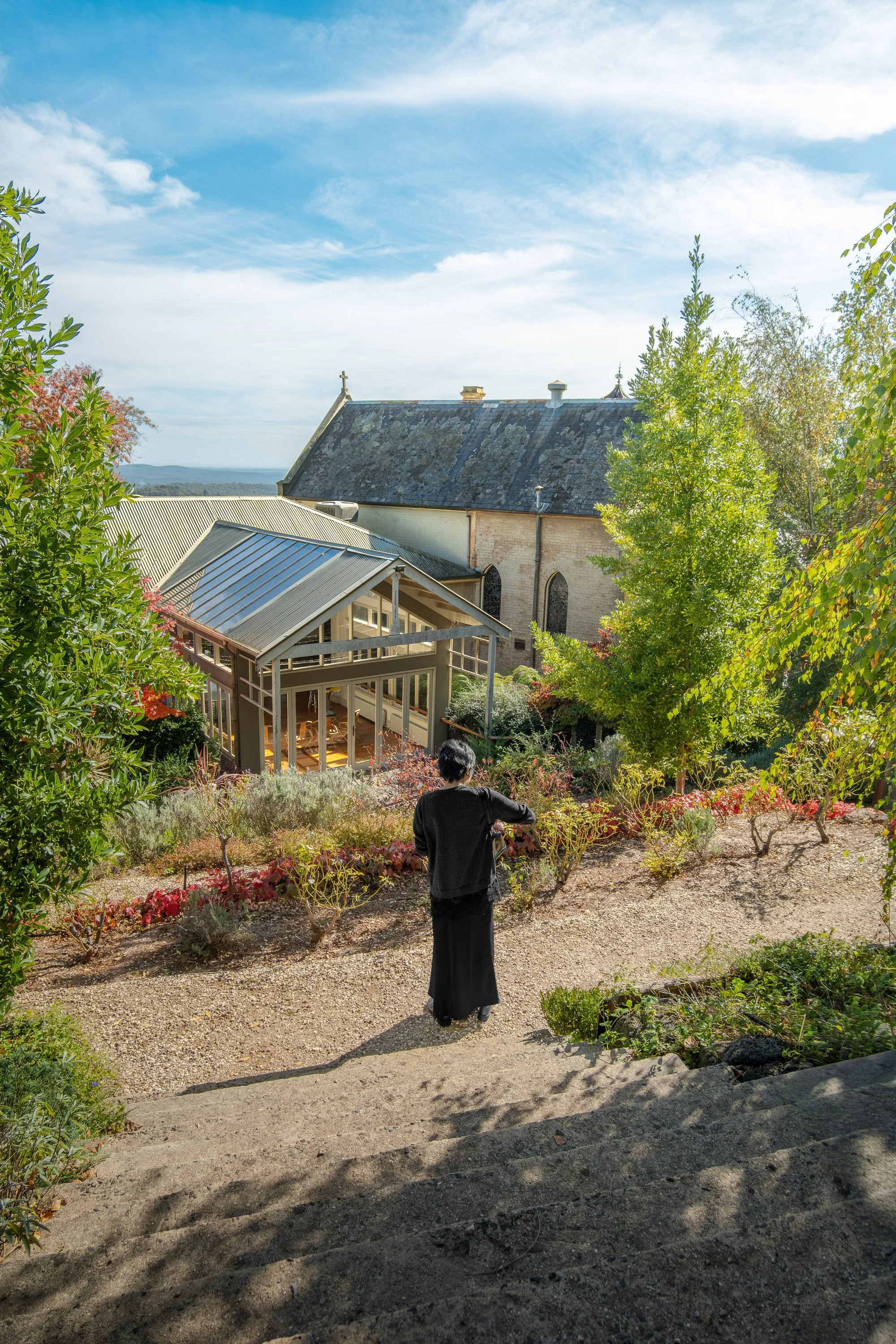 A person dressed in black walking down a set of stone steps in a garden with trees and shrubs, with a church building with a sloped roof and a stone exterior in the background under a partly cloudy sky.