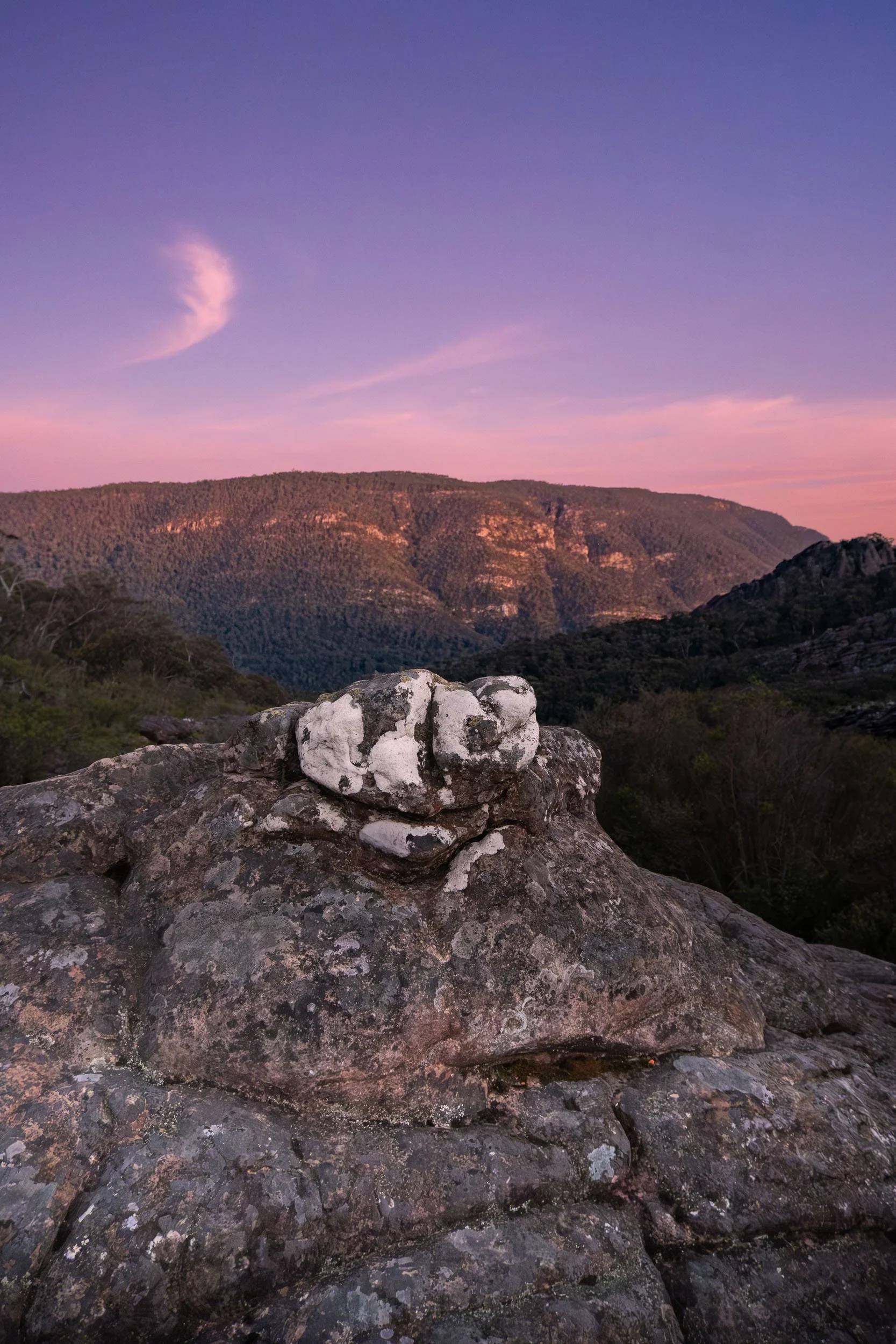 A landscape view of rolling hills under a purple sunset sky with scattered clouds, rocky foreground with lichen-covered stones.