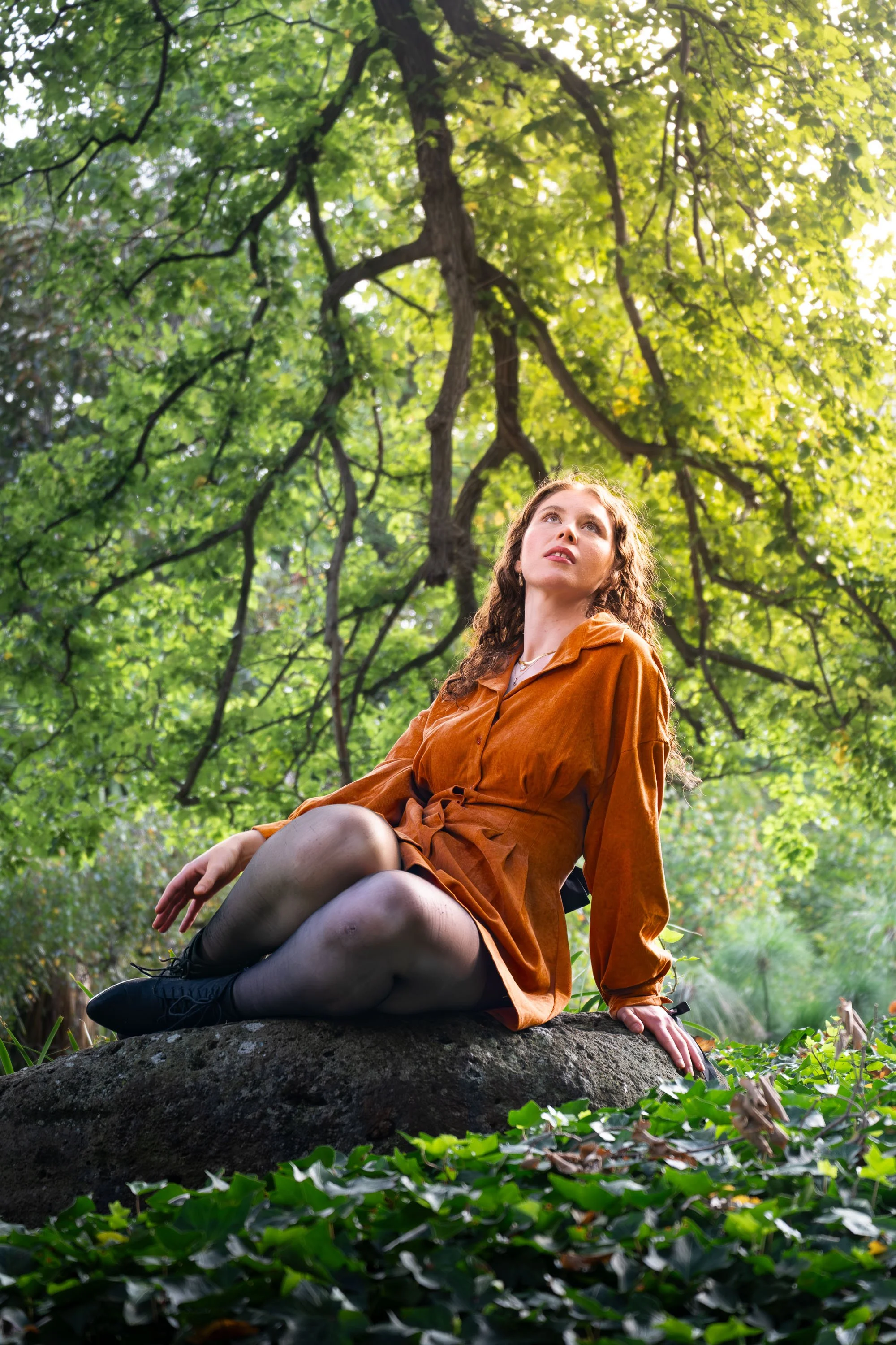 A woman with curly hair, wearing a rust-colored dress and black shoes, seated on a large rock in a lush green forest, looking upward toward the sunlight filtering through the trees.