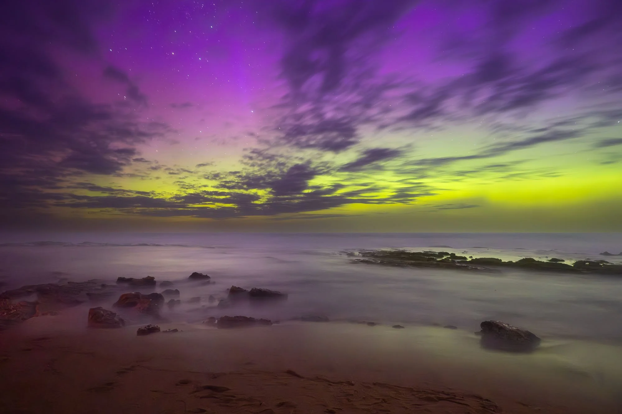 A long exposure photograph of a night sky over the ocean with colorful auroras in shades of purple, green, and yellow, with rocks on the shoreline in the foreground.