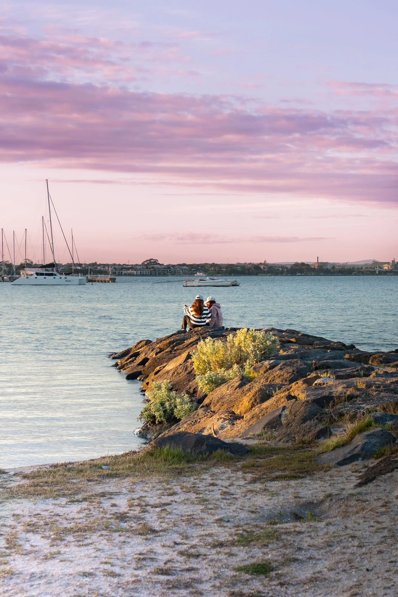 A coastal scene at sunset with two people sitting on rocks by the water, boats anchored in the distance, and a sky with pink and purple clouds.