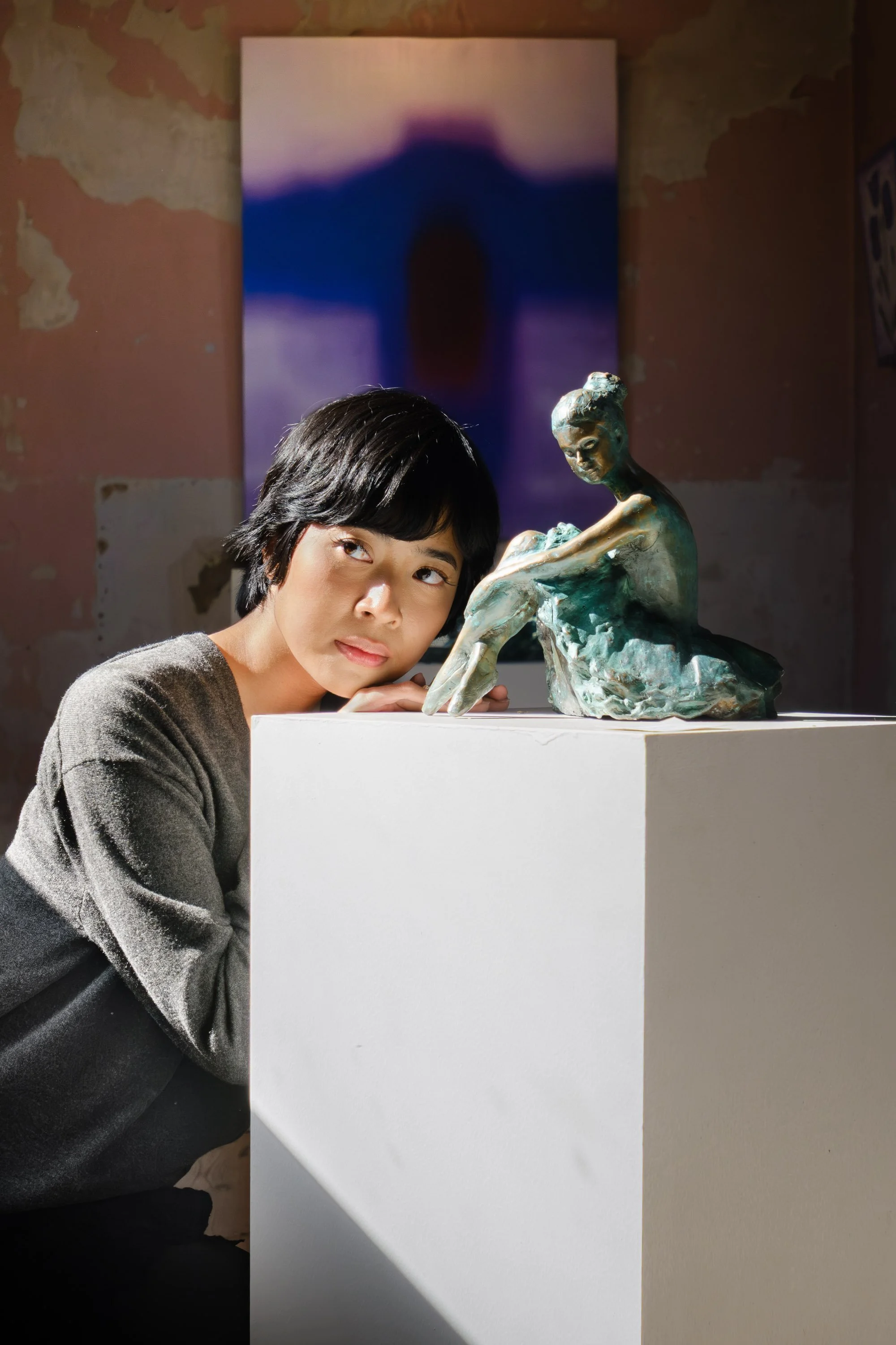 A woman with short black hair resting her head on her hand next to a bronze sculpture of a seated woman in a gallery space. There is an abstract painting with a dark shape in the background, and the wall behind the woman and sculpture appears worn an