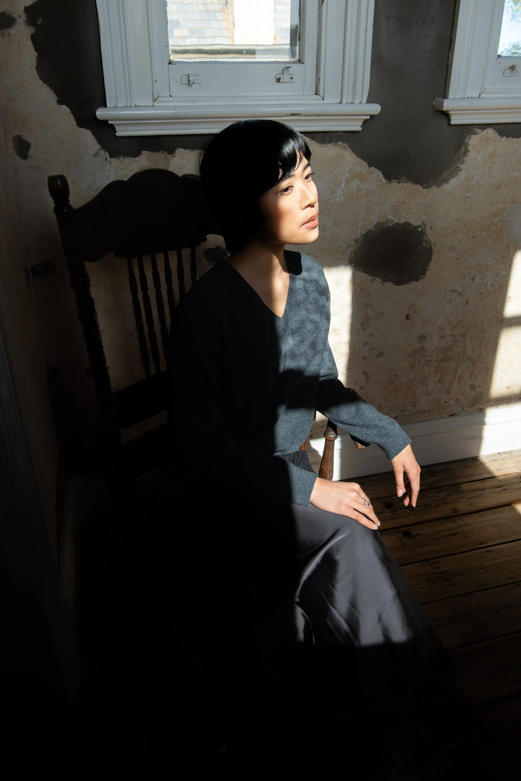 A woman with short black hair sitting on a wooden floor next to a wall with peeling paint, illuminated by sunlight through a window.