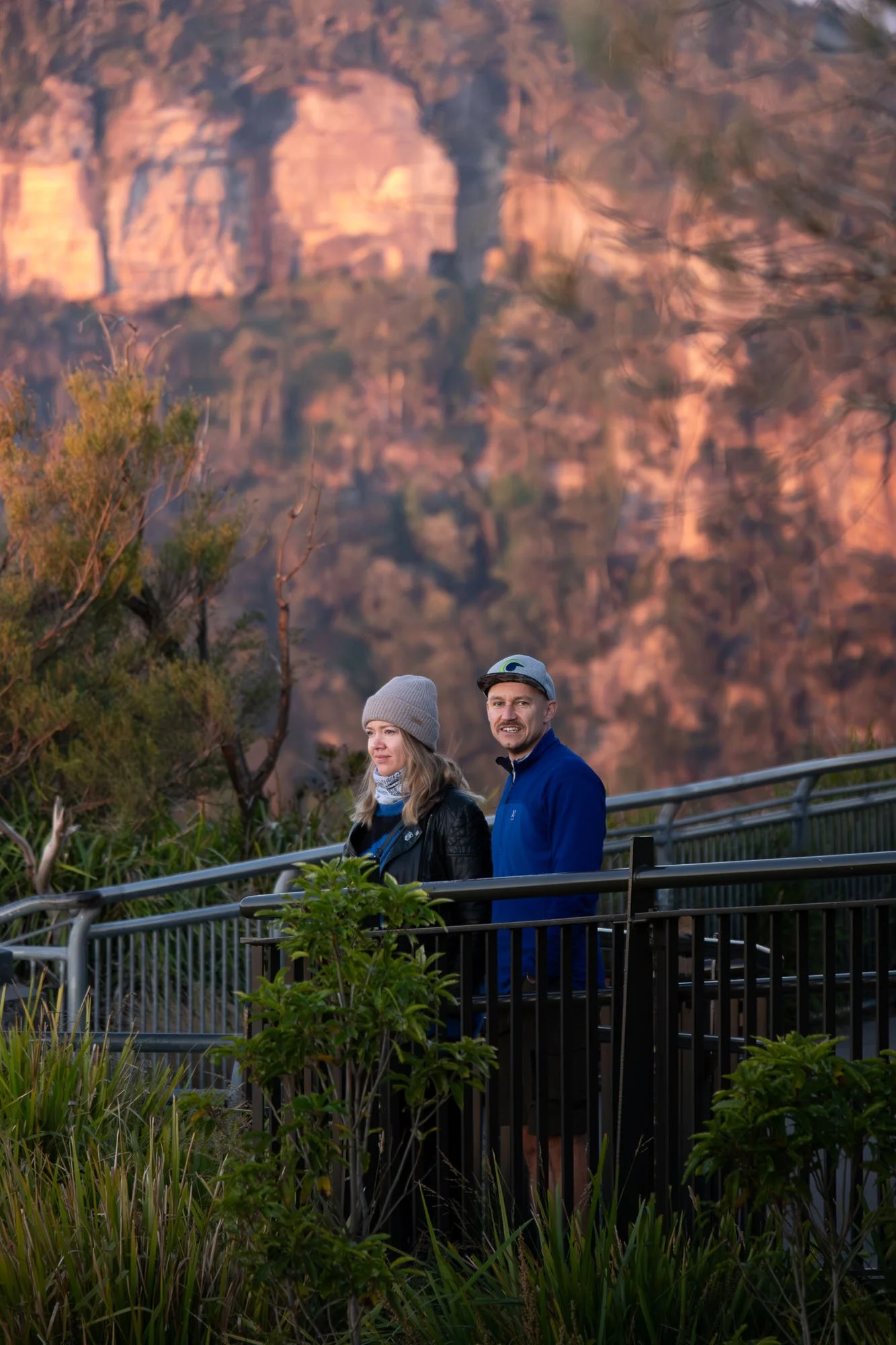 Two people standing on a viewing platform with a mountain landscape in the background, one woman and one man dressed warmly, in the early morning or late afternoon light.