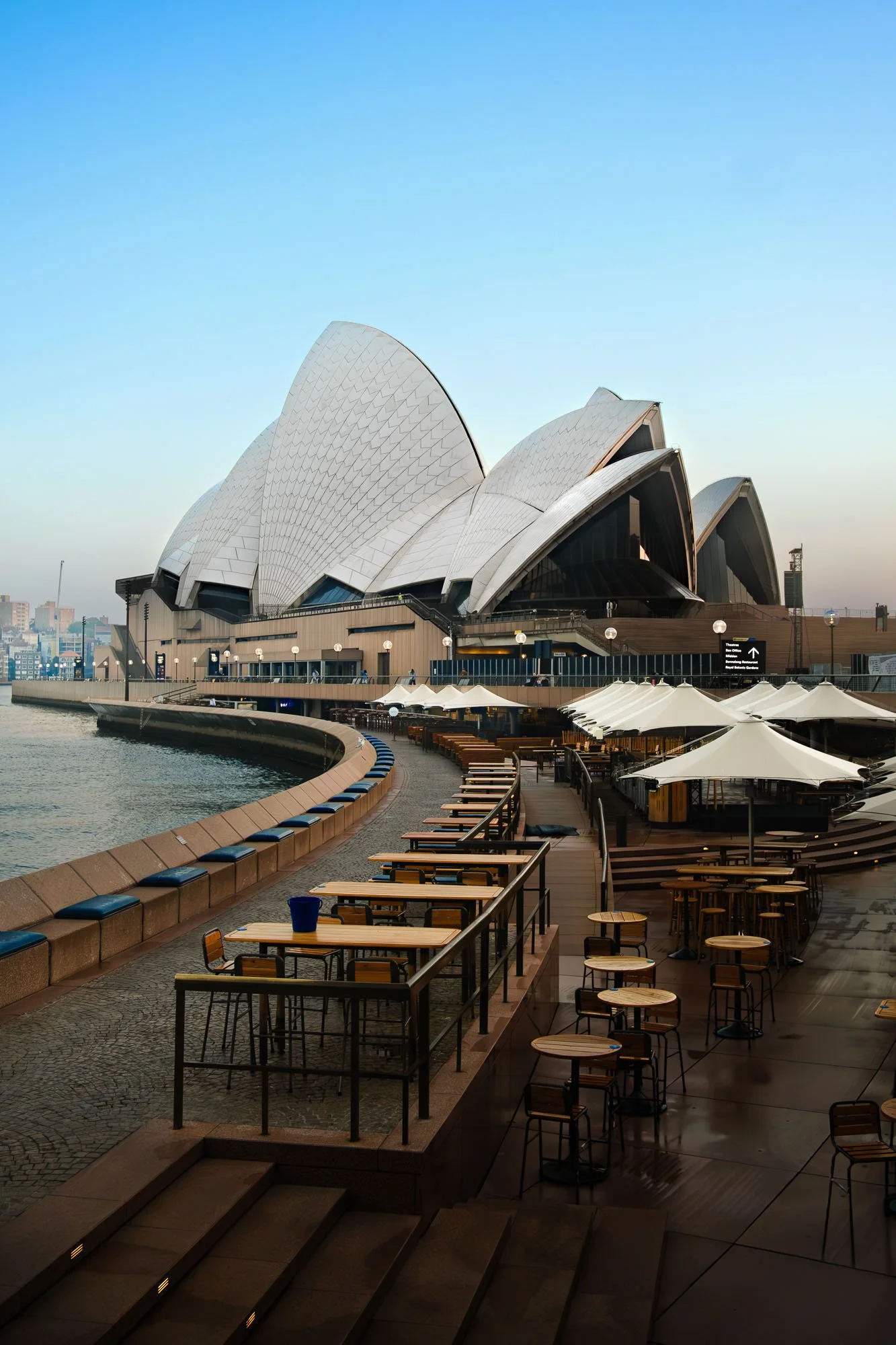 Sydney Opera House with waterfront seating and umbrellas in the foreground during early morning or late evening.