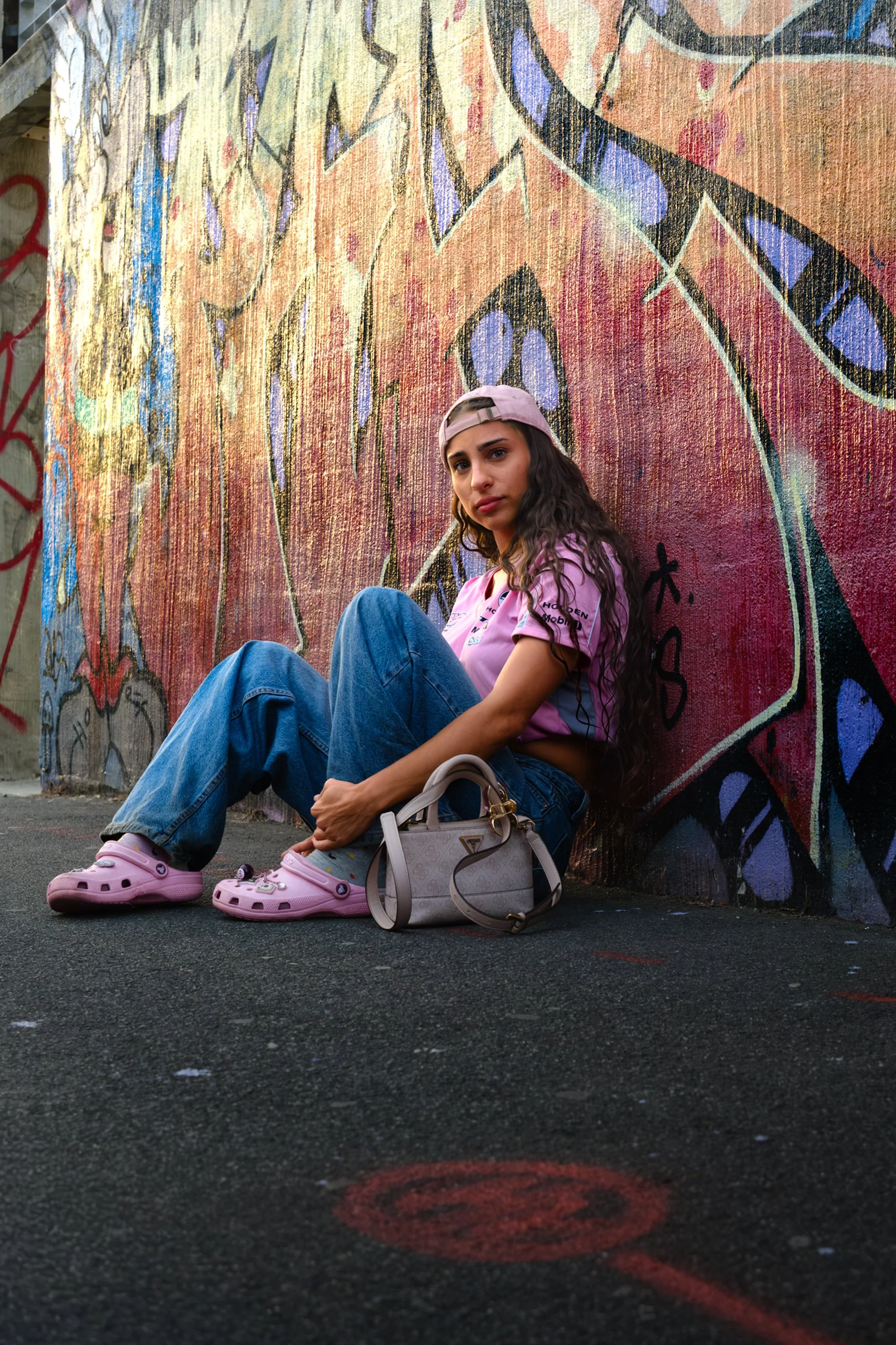 A young woman with long curly hair, wearing a pink baseball cap backwards, pink t-shirt, ripped jeans, and pink Crocs, sitting against a colorful graffiti wall on a dark street. She has a beige handbag placed beside her and looks at the camera with a
