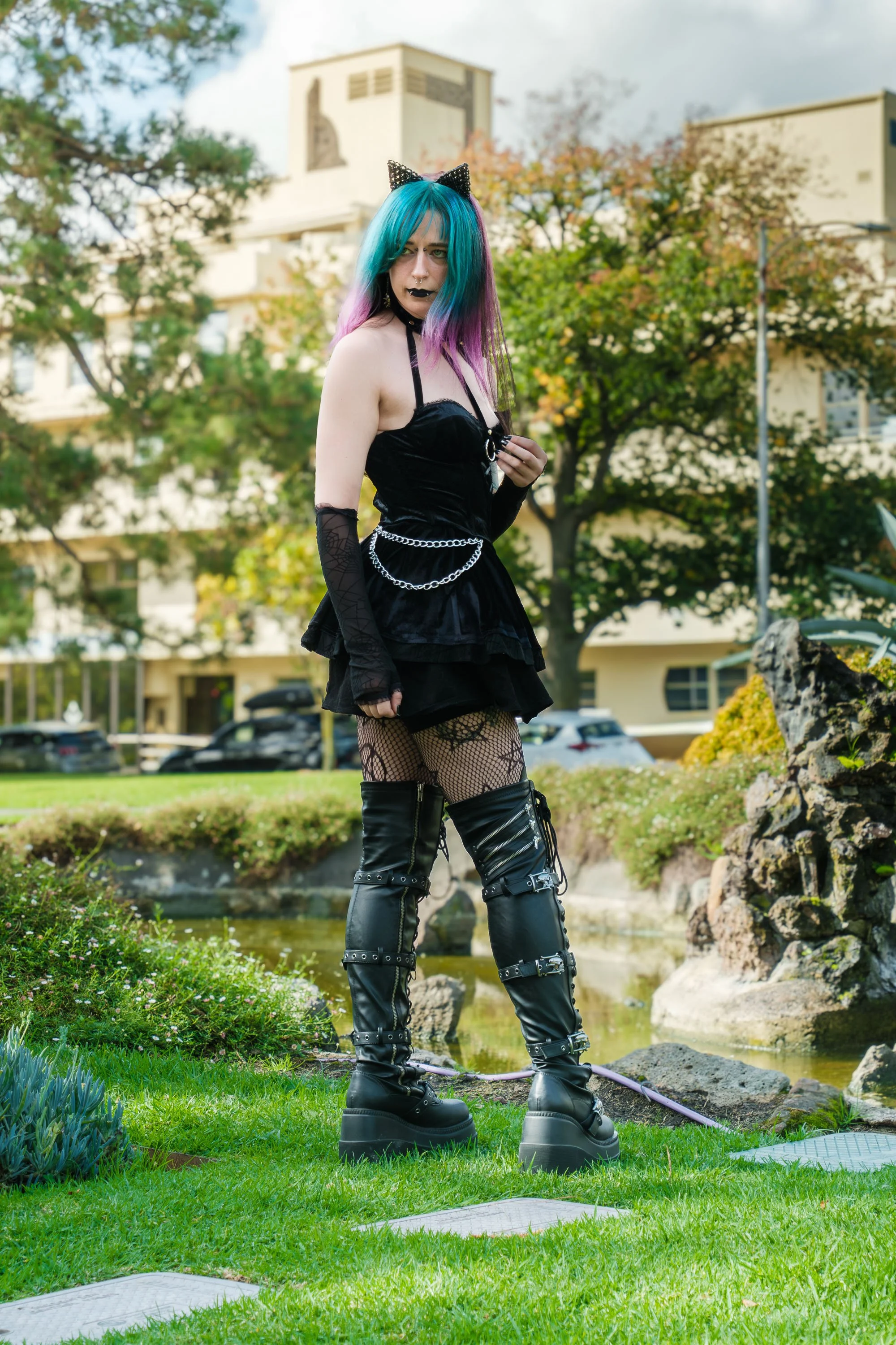 Young woman with rainbow-colored hair, wearing black gothic-style clothing and platform boots, standing outdoors by a pond with rocks and greenery in the background.