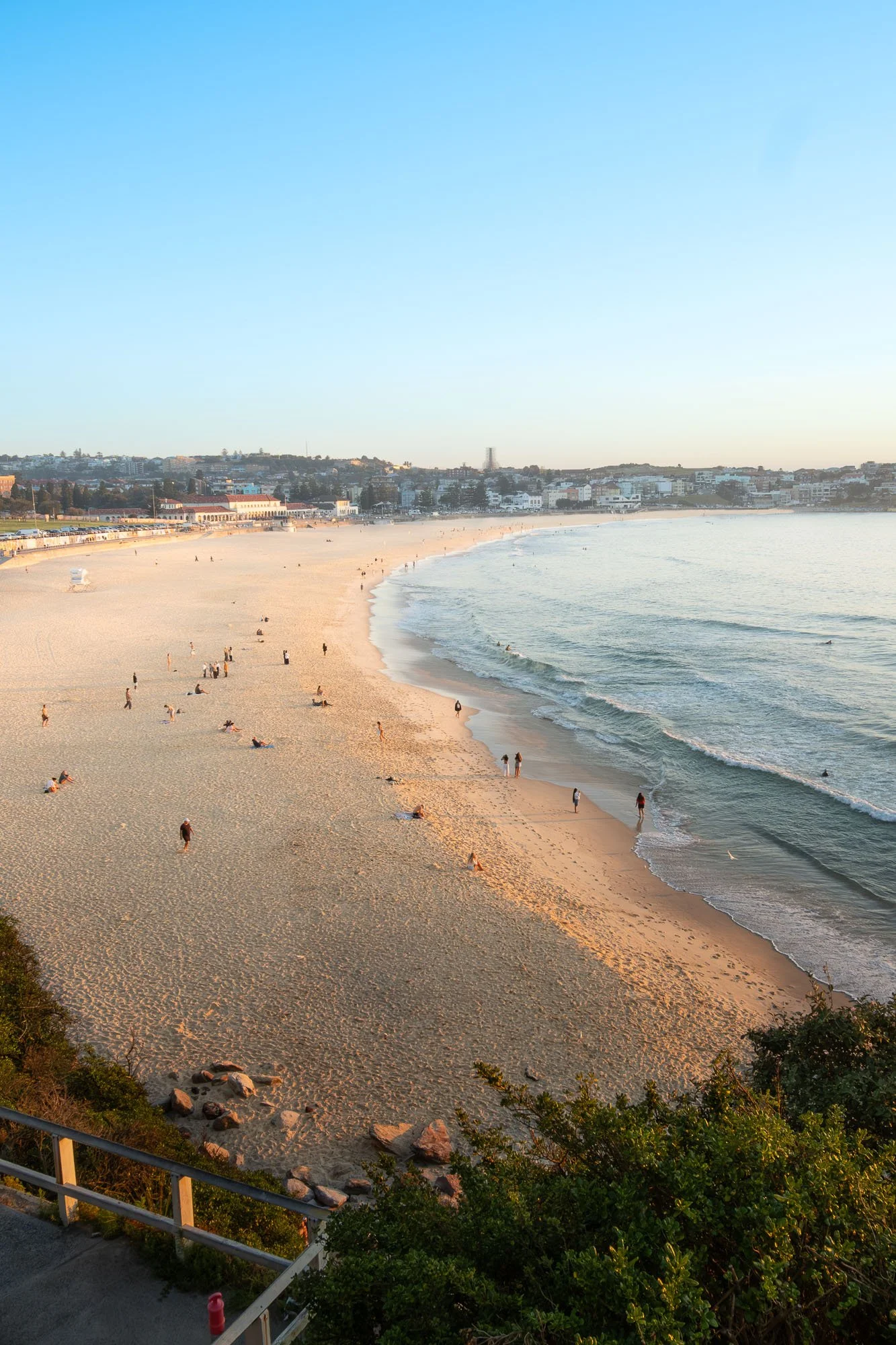 A sandy beach with people walking and relaxing, waves gently lapping the shore, and a cityscape in the background with buildings and houses under a clear blue sky.