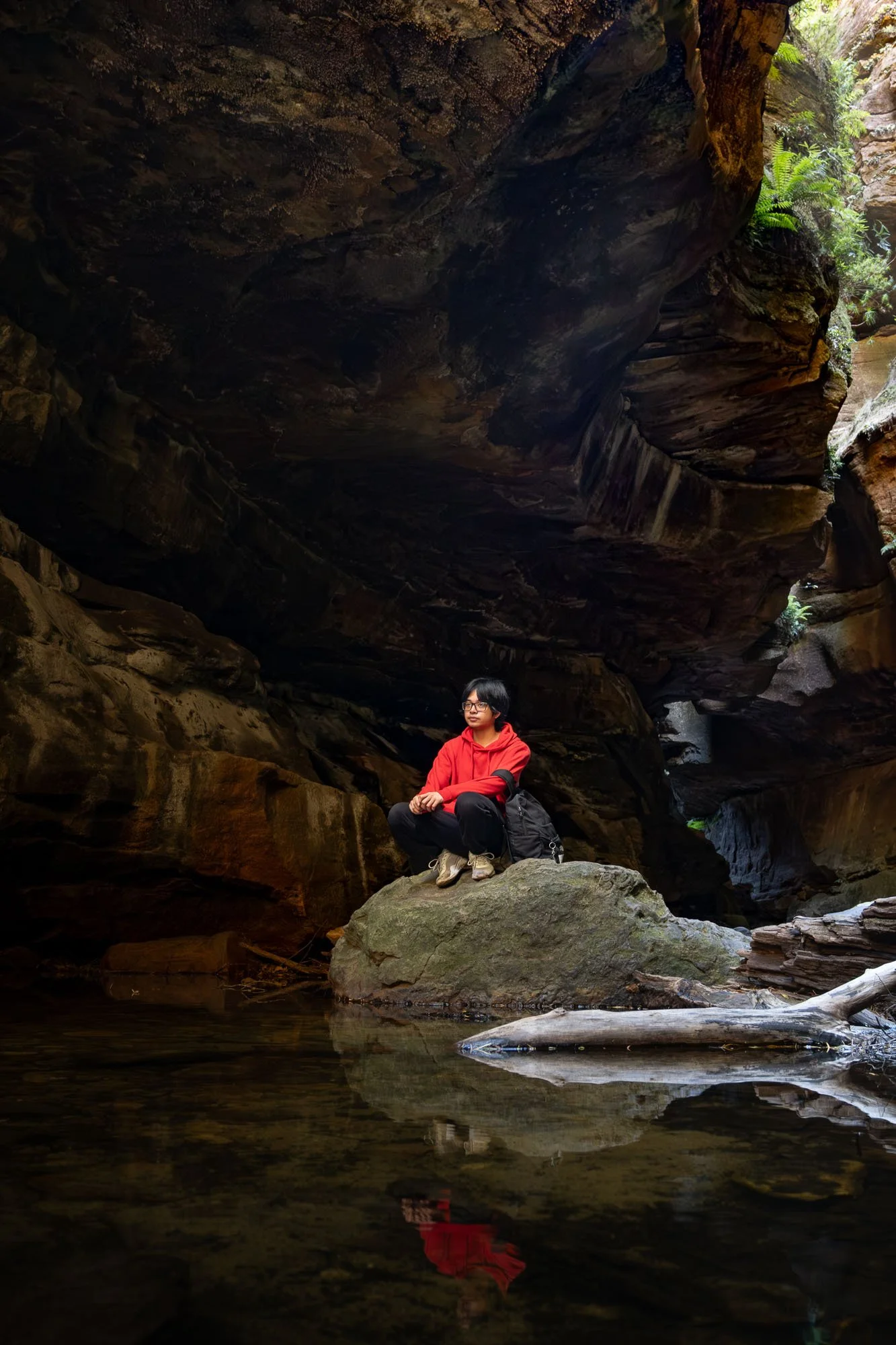 A person wearing a red hoodie and glasses is sitting on a rock in a canyon, with lush green plants on the rocks and a stream at their feet. The rocky walls of the canyon tower above and surround the person.