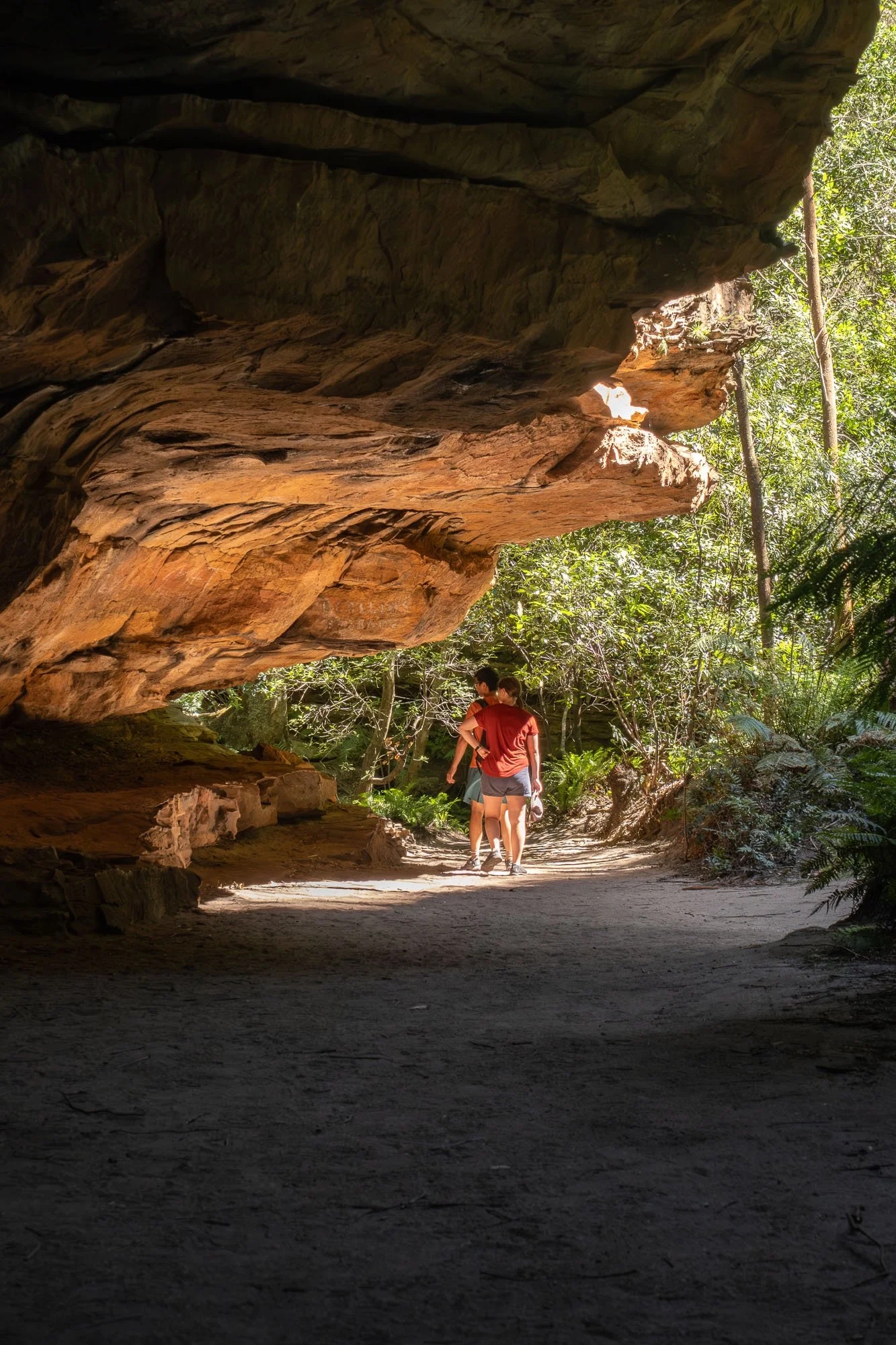 People walking underneath a large overhanging rock formation in a forested area.