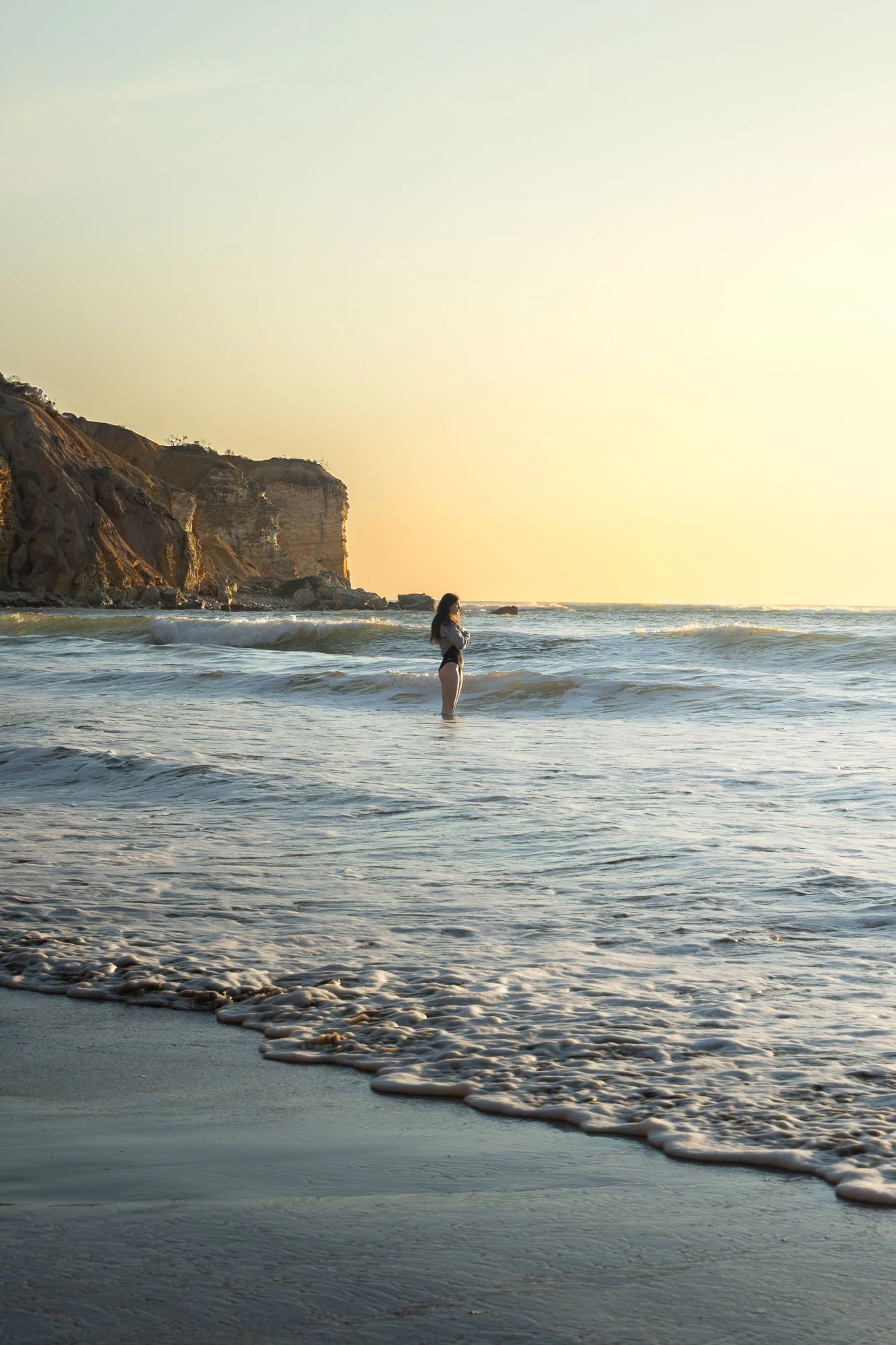 A woman stands in shallow ocean water near a rocky cliff during sunset.