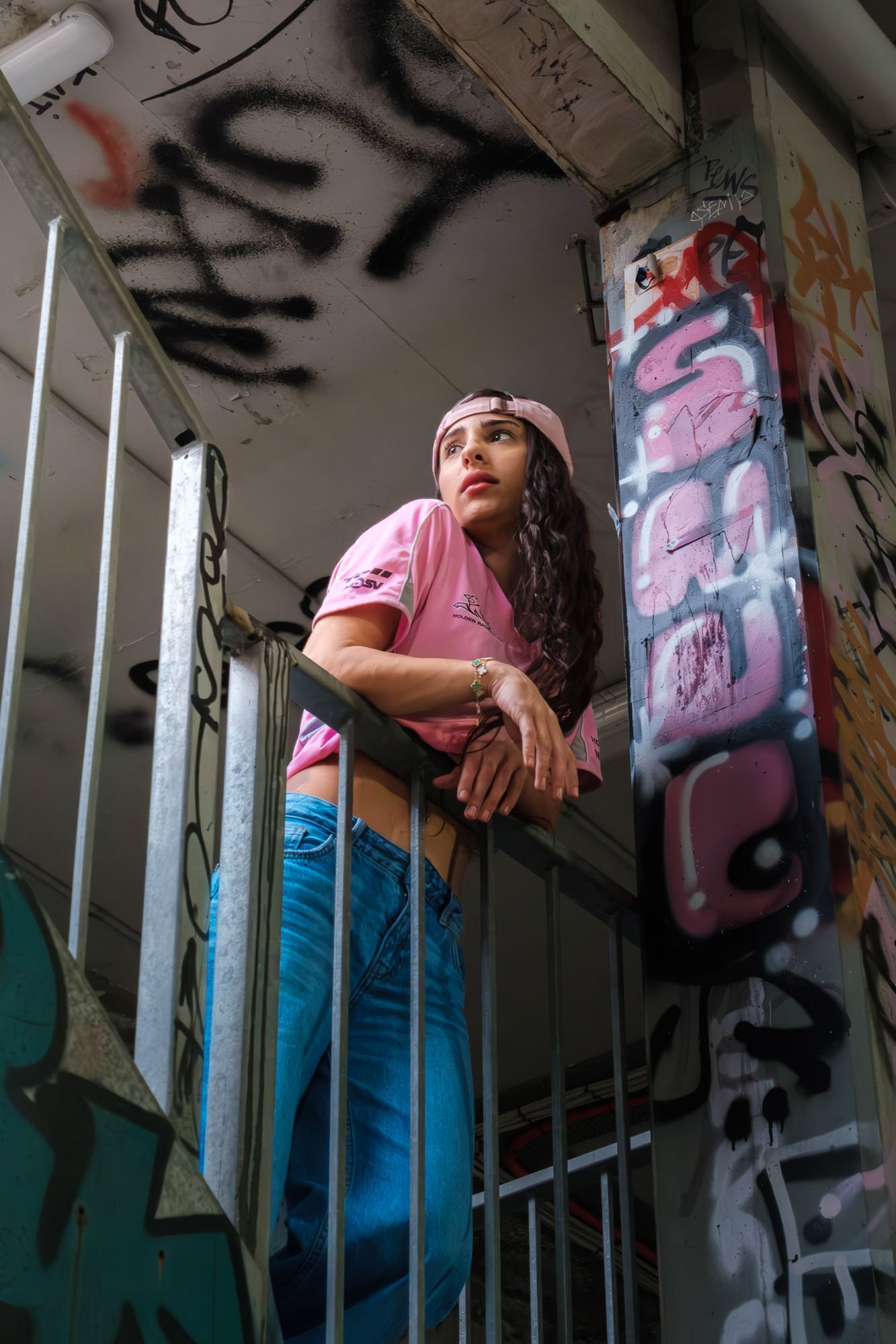 A young woman with long curly hair, wearing a pink baseball cap and a pink sports jersey, leans on a metal railing in front of graffiti-covered walls and ceiling, looking upward with a thoughtful expression.