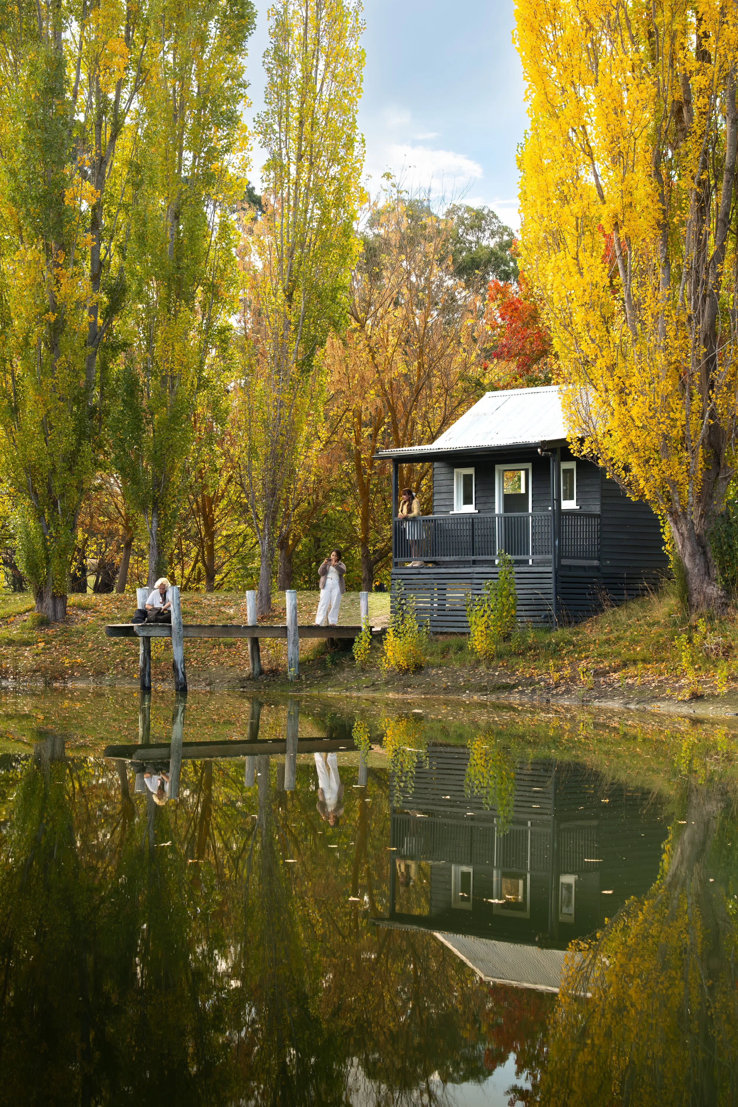 A black cabin with a silver roof on a small hill next to a body of water surrounded by tall trees with yellow and green leaves in autumn, reflecting in the water, with a woman in white standing on a dock and a person sitting on a bench