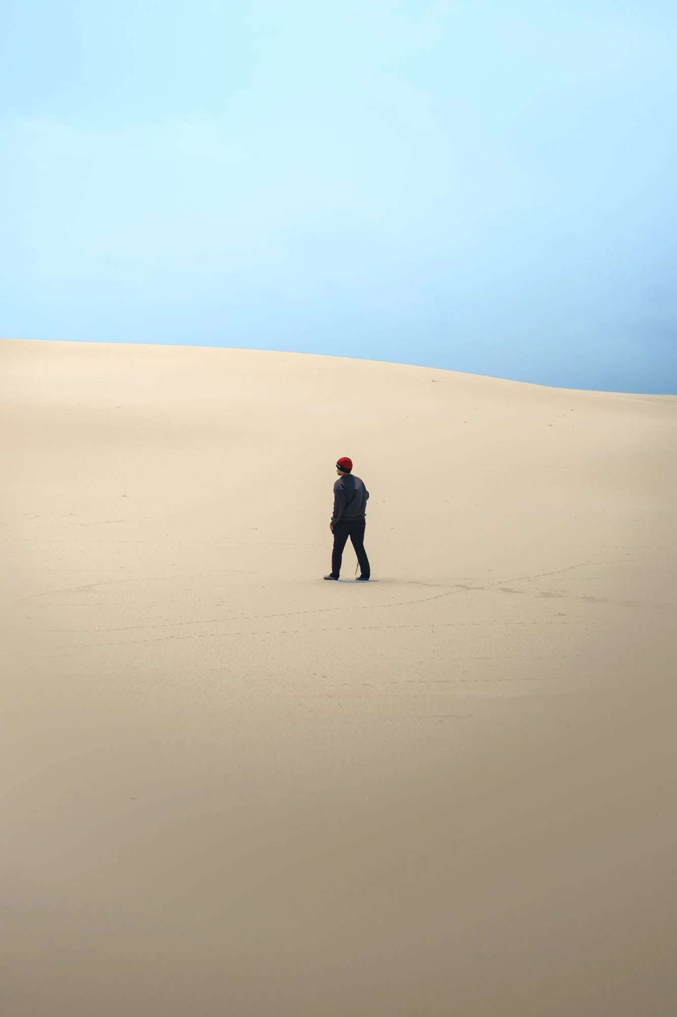 A person wearing a red beanie walking on a sandy desert landscape with a blue sky overhead.