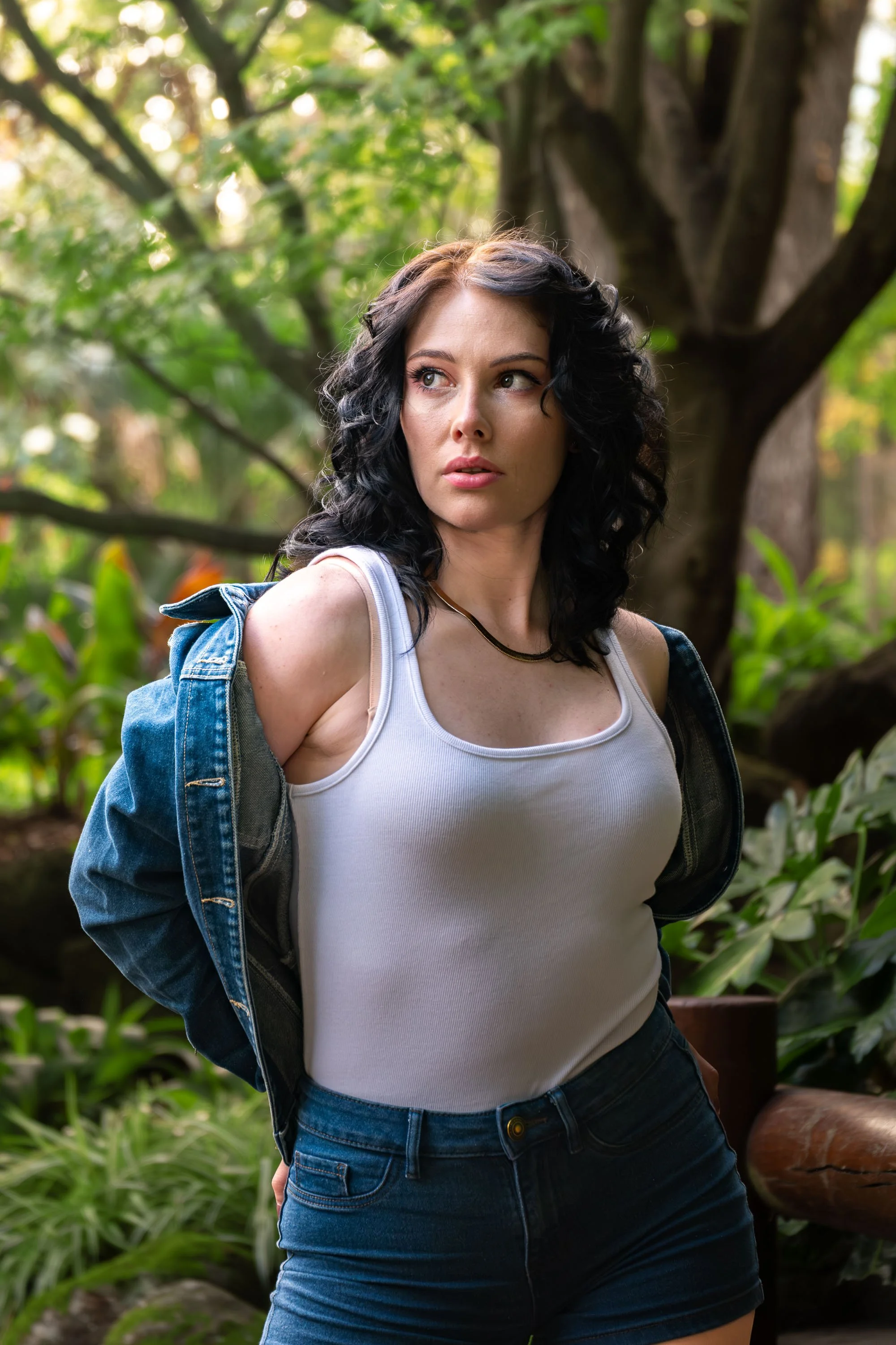 A woman with black curly hair and fair skin looking to the side, outdoors surrounded by trees and green foliage, wearing a white tank top, a black denim jacket off her shoulders, and dark jeans.