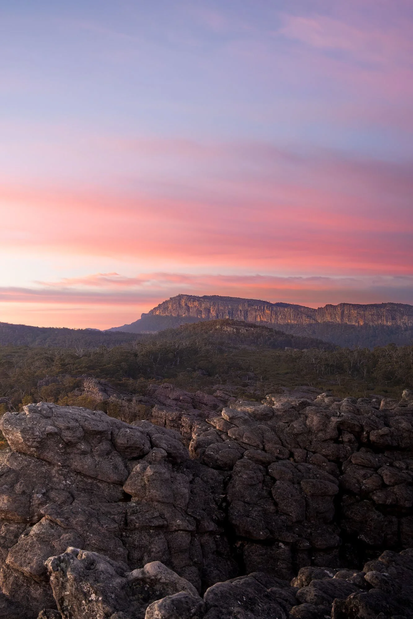 A landscape featuring a rocky foreground, a range of trees and a plateau in the background under a colorful sunset sky with pink, purple, and blue hues.