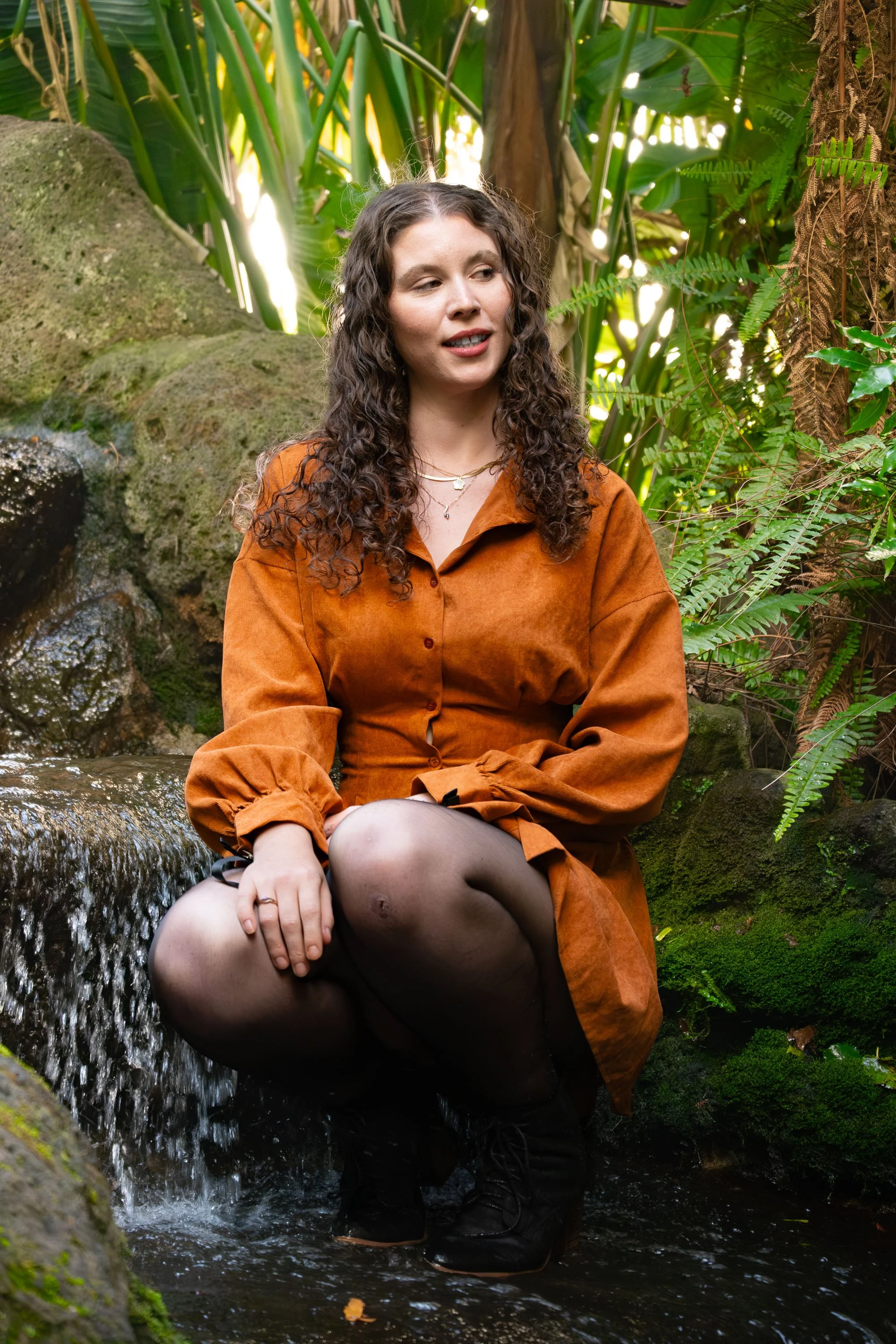 A young woman with curly brown hair sitting in a small stream surrounded by lush greenery and moss-covered rocks, wearing an orange dress and black boots.